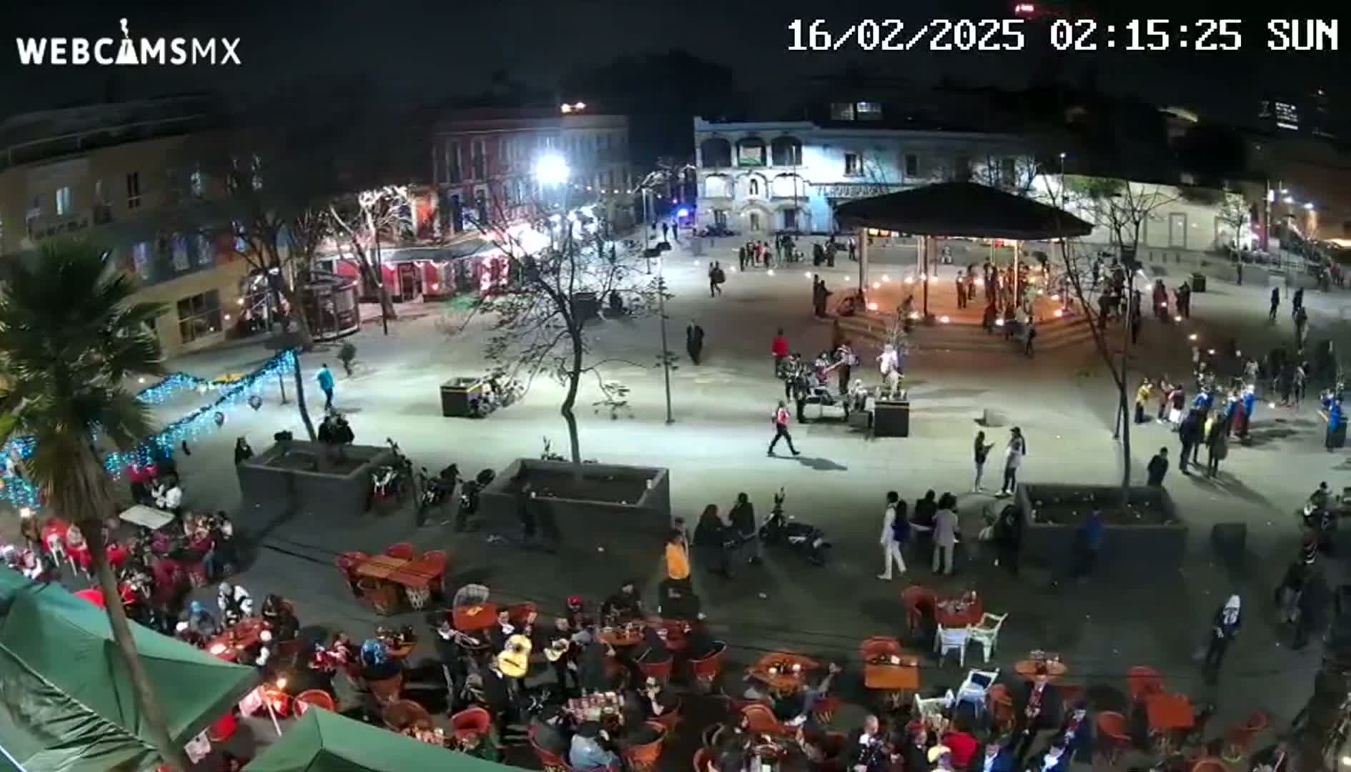A nighttime, outdoor scene shows a town square filled with people gathered around tables at cafes and a central gazebo, with string lights illuminating the area.