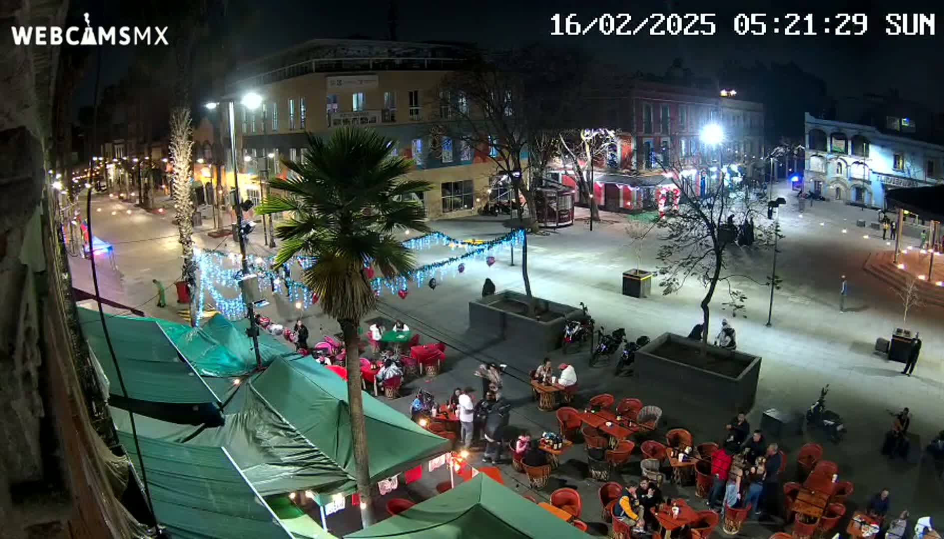 A nighttime view of a town square with several people sitting at outdoor tables under market tents, and a streetcar visible in the background under clear skies.