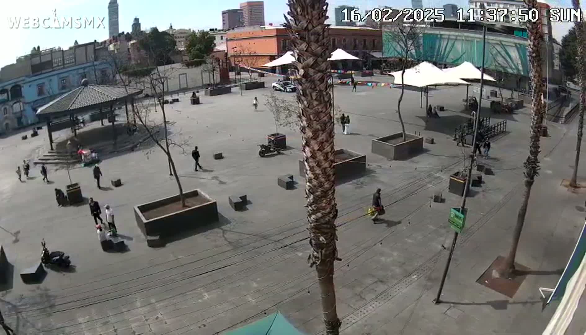 A mostly empty plaza with a few people walking around, some potted plants, and a gazebo under a sunny sky.