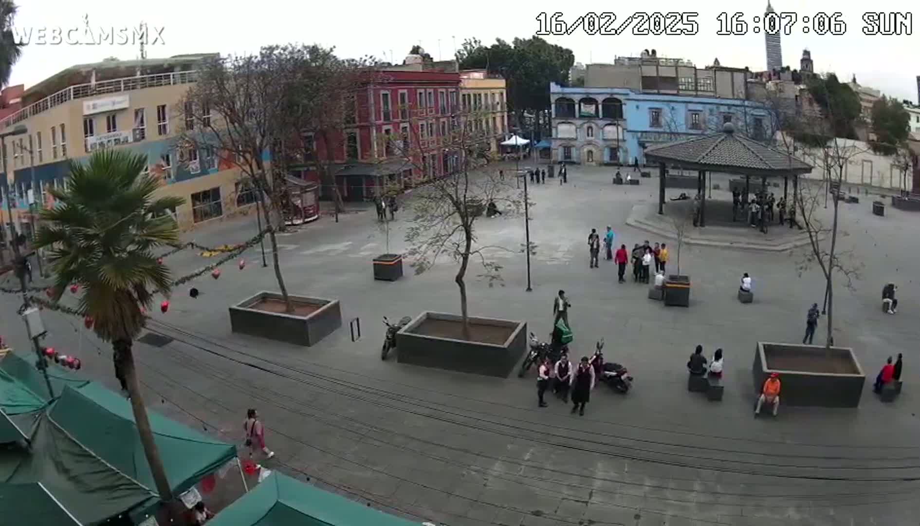 A mostly empty town square with several buildings, a gazebo, and some people, under an overcast sky.