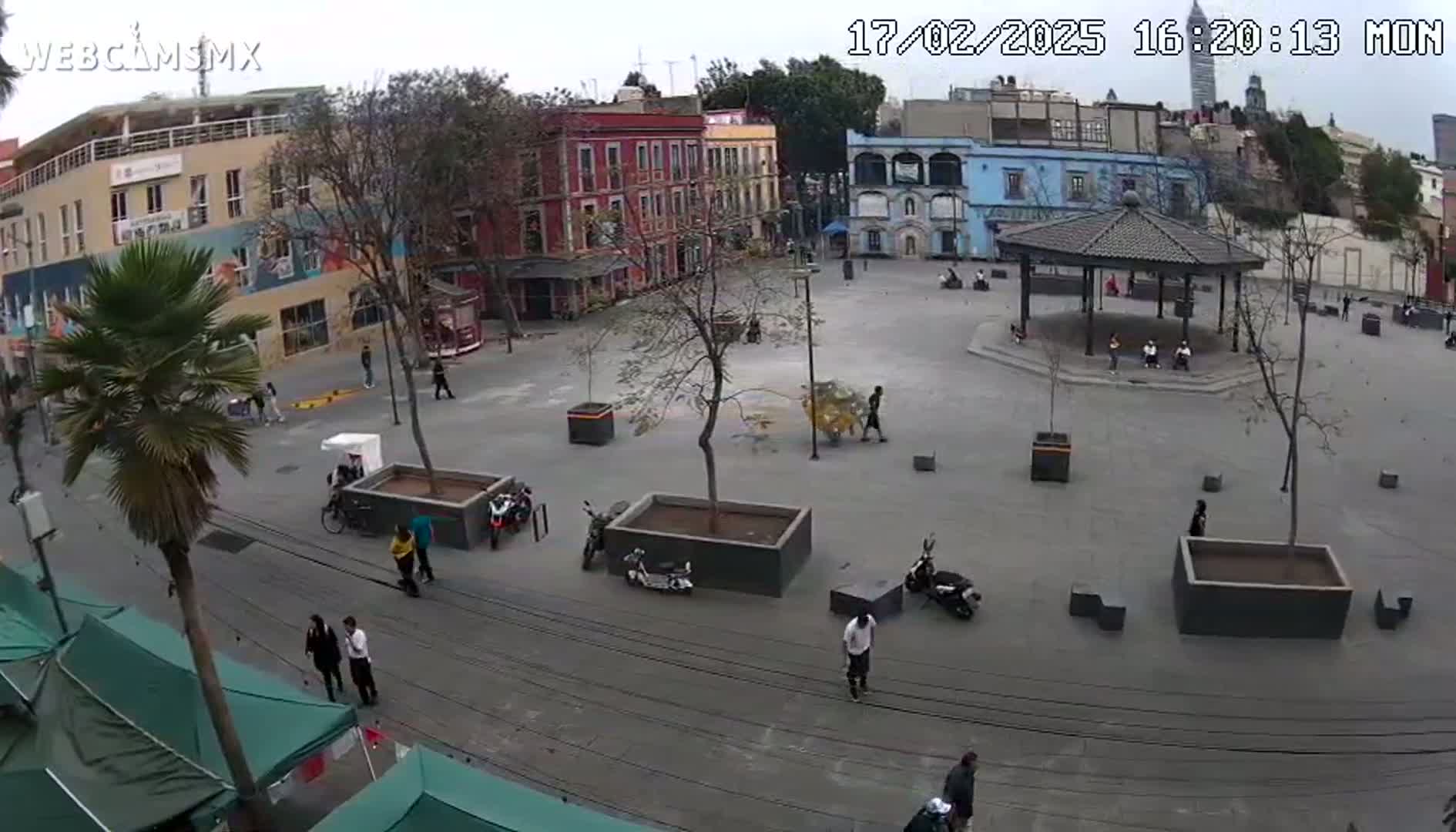 A mostly empty plaza with several buildings surrounding it, under a cloudy sky, features people walking around, some parked scooters, and a gazebo in the center.