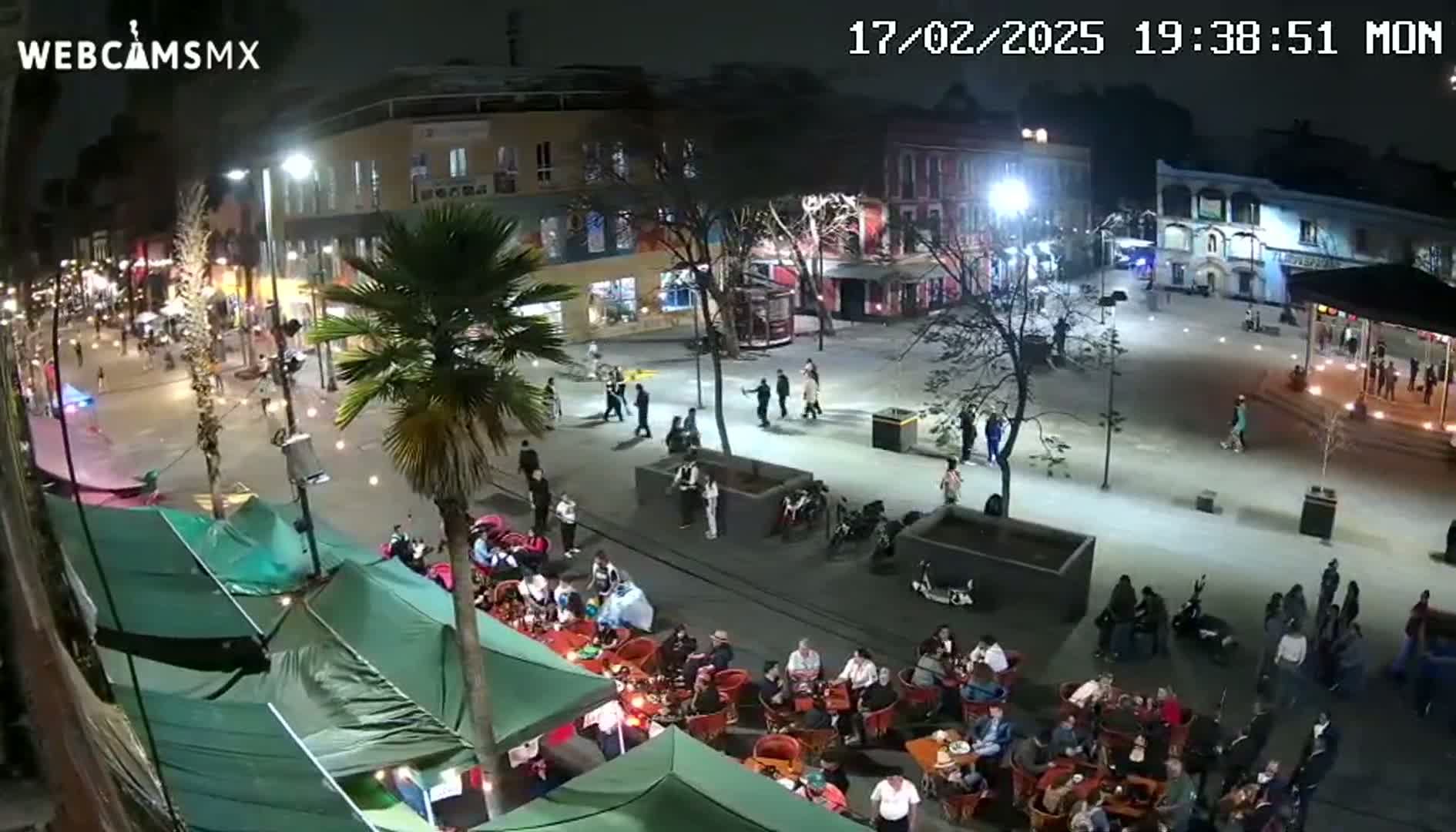 A nighttime, brightly lit plaza and street scene shows people walking and sitting at outdoor cafes under a clear sky.