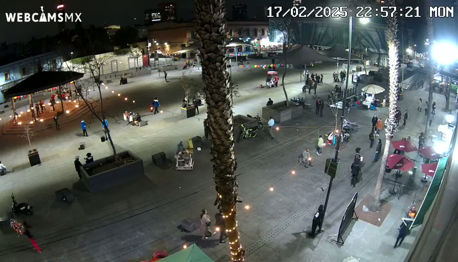 A nighttime aerial view of a plaza with people walking and gathering around various areas, illuminated by string lights and building lights.
