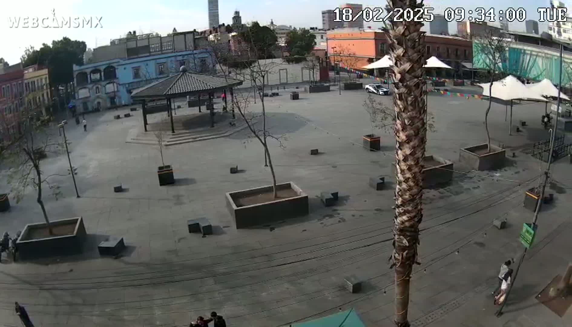 A mostly empty plaza with a gazebo and several trees is seen on a partly sunny day, with a few people scattered about.