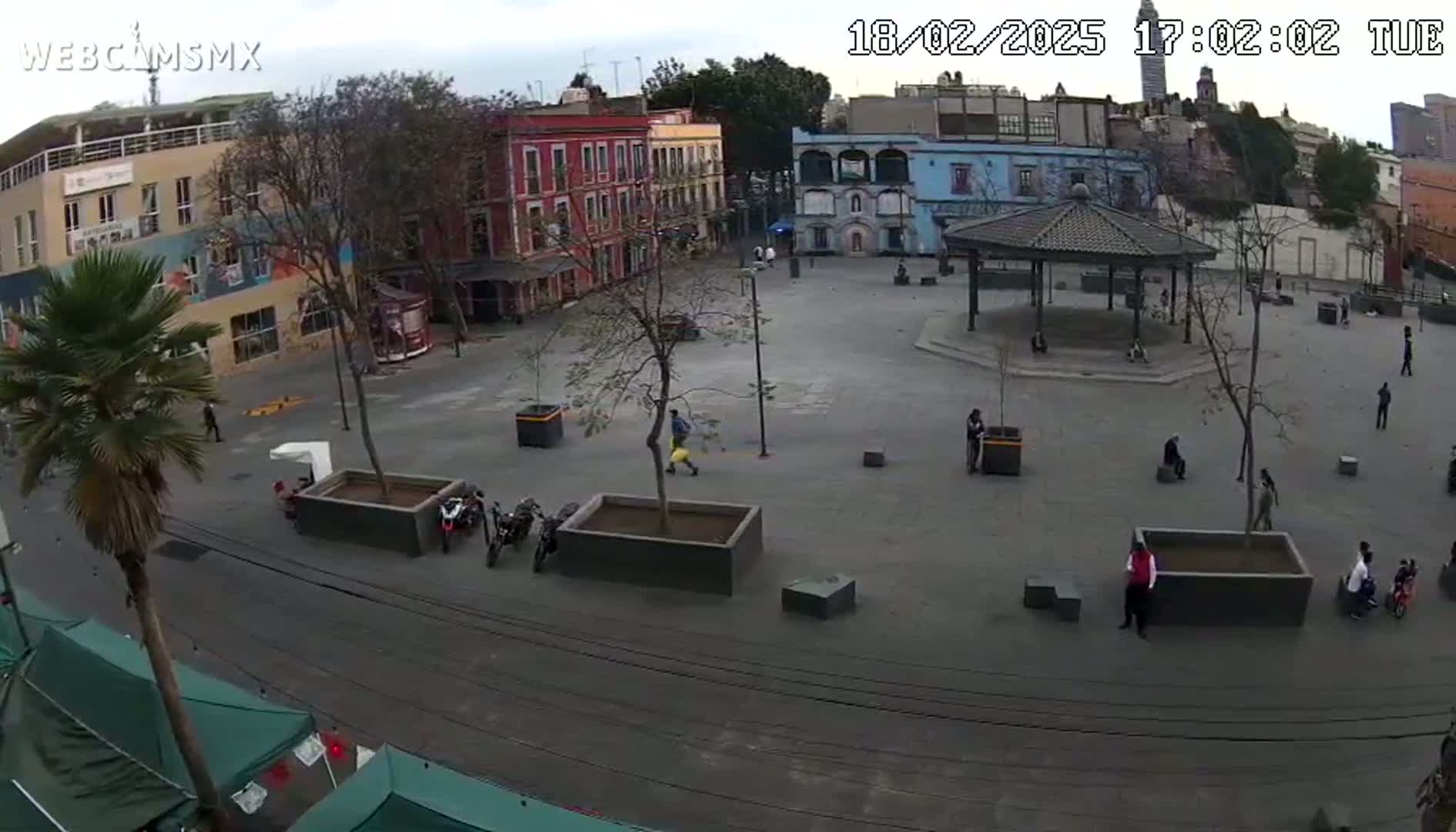 An overcast day shows a plaza with several people scattered throughout, some parked motorcycles, and buildings surrounding the area.