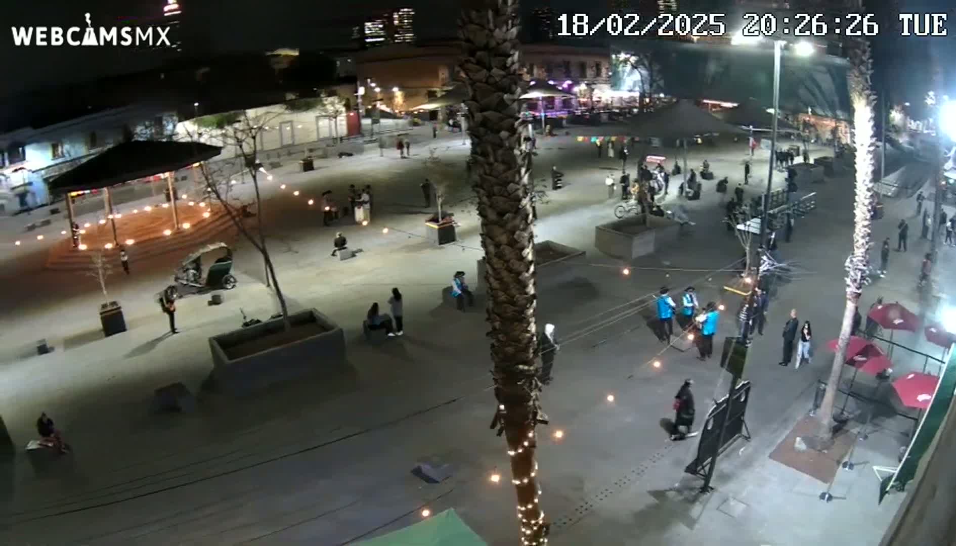 An outdoor plaza at night is sparsely populated with people, illuminated by string lights and ambient lighting, under clear skies.