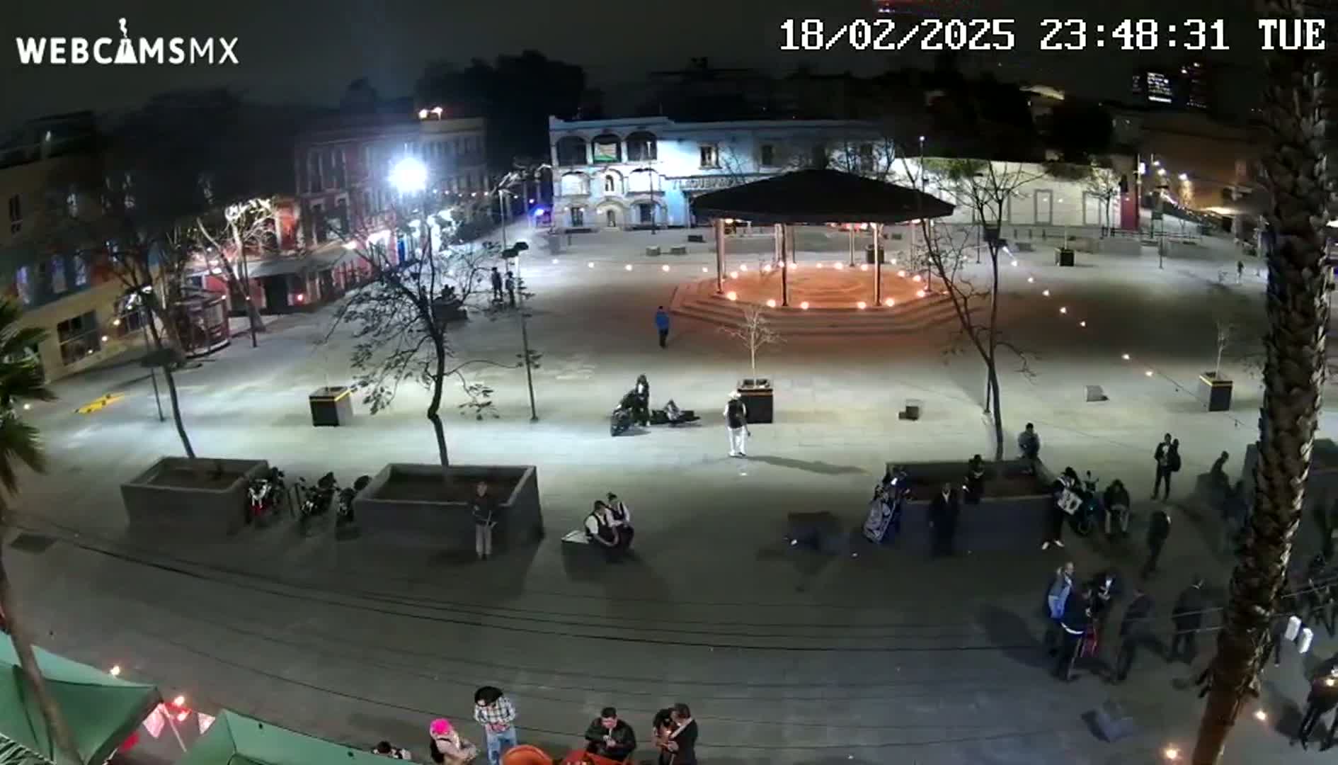 A nighttime, brightly lit plaza with people gathered in small groups, some seated, others standing, near a gazebo and surrounding buildings.