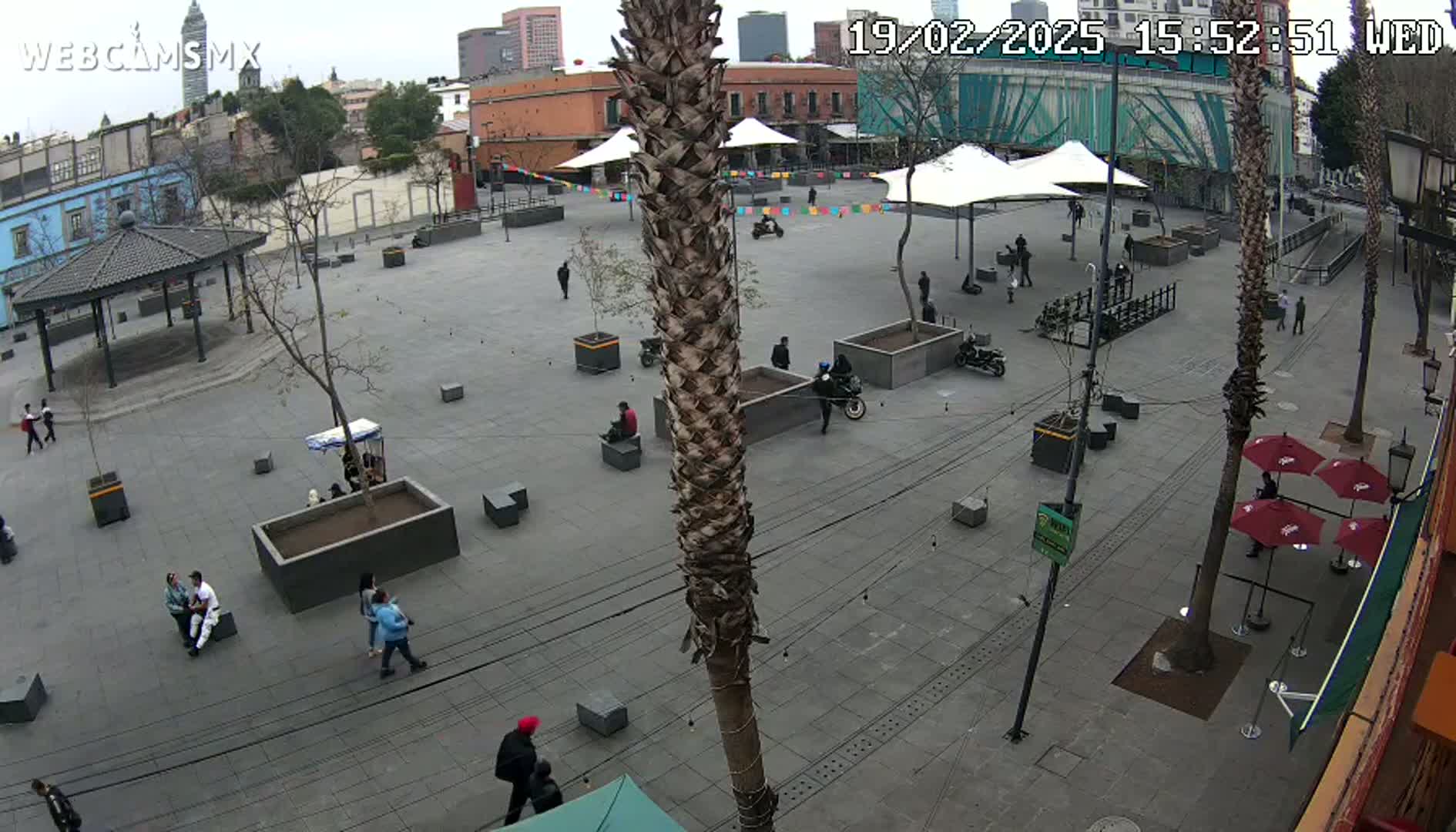 A mostly empty plaza with several people scattered about, some seated at tables under umbrellas, is viewed from above on an overcast day.