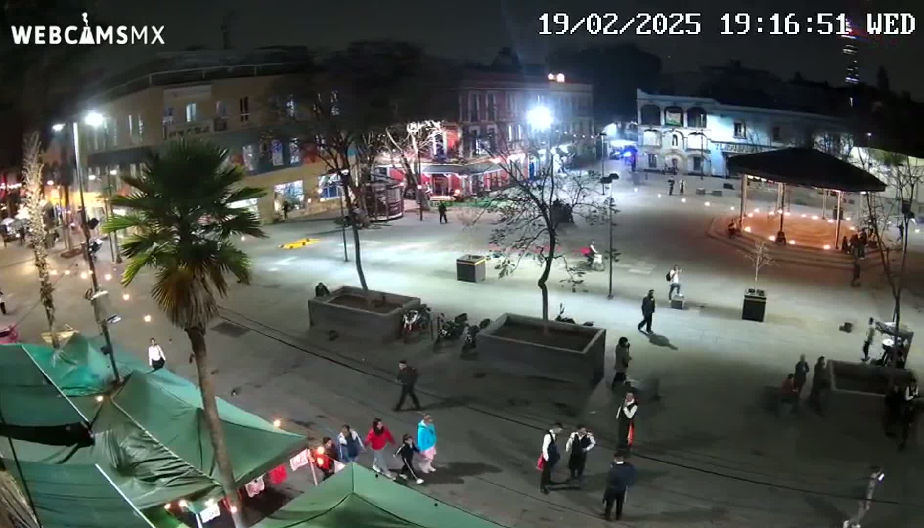 A nighttime, brightly lit town square with people walking around, some vendors selling goods from tents, and a gazebo in the center.