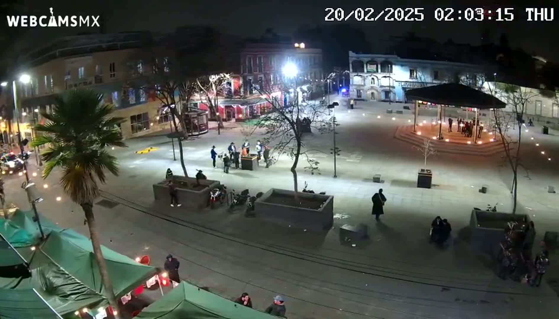 A nighttime, brightly lit plaza scene shows several people gathered in small groups, some near a gazebo, others near street-side stalls, with motorcycles parked nearby and a streetcar visible in the background.