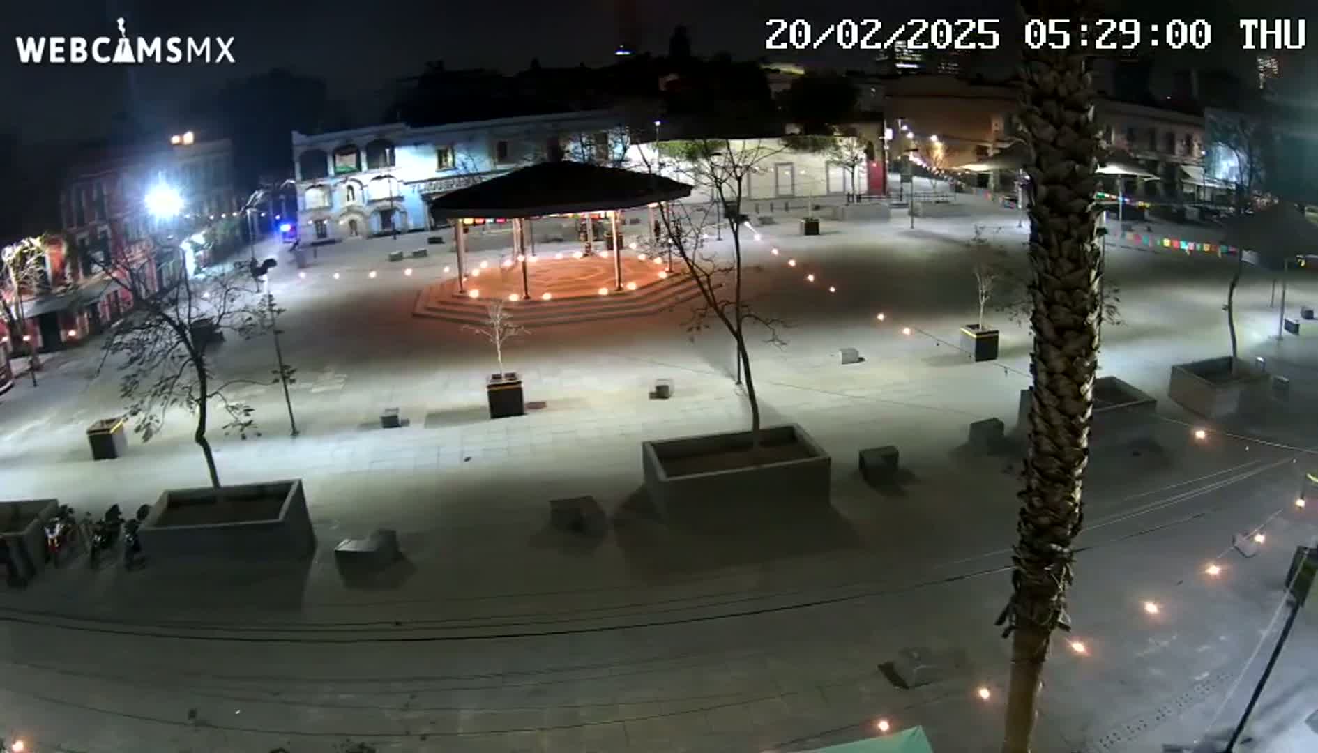 A nighttime aerial view of a plaza with a gazebo at its center, surrounded by buildings and illuminated by lights.