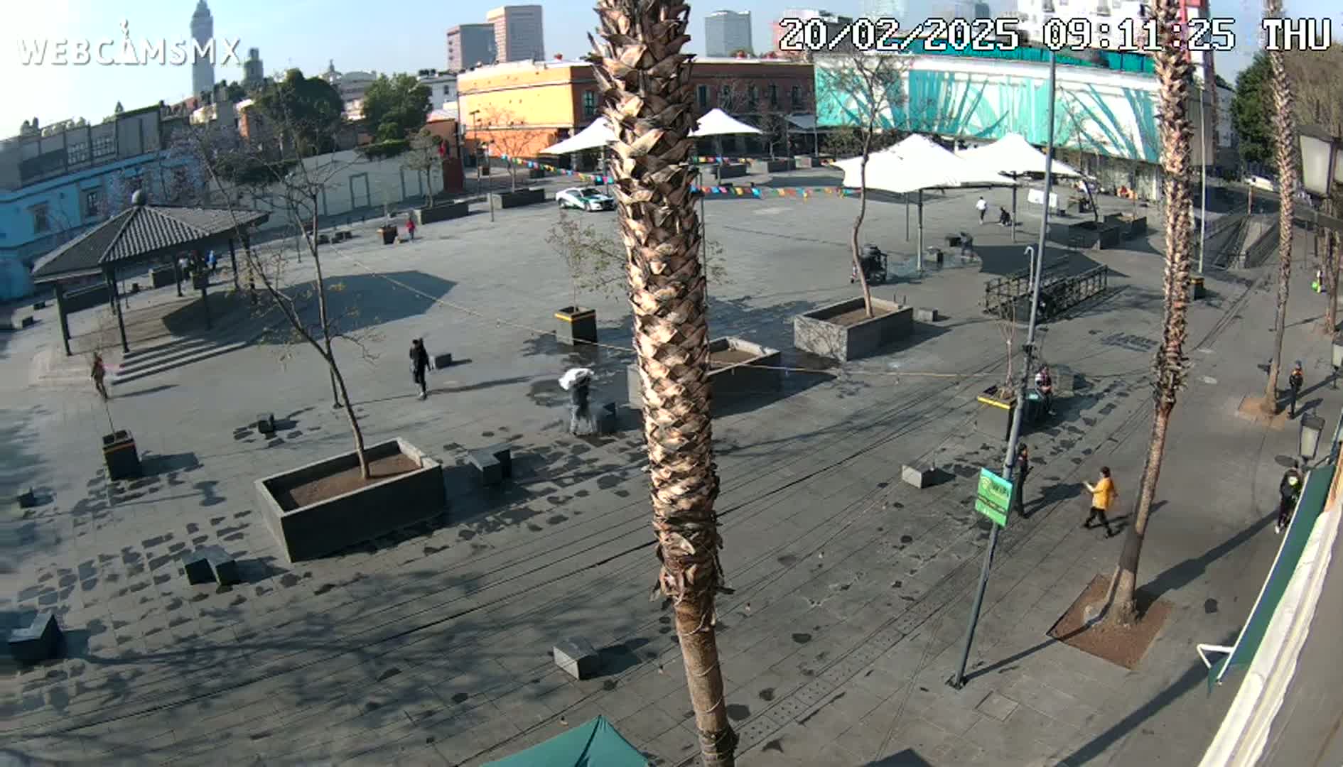A mostly empty plaza with a few pedestrians, several palm trees, and a gazebo under a sunny sky.