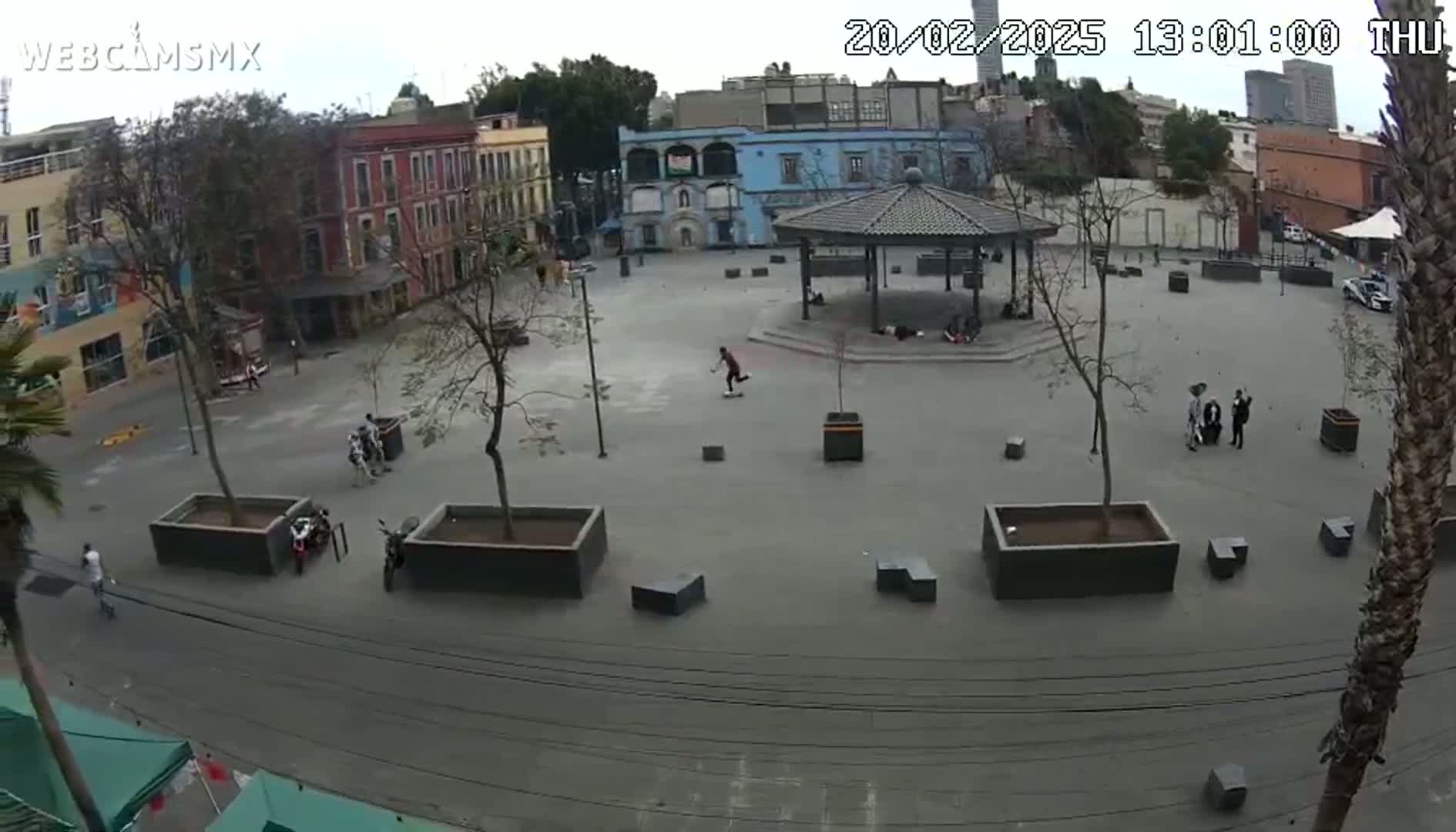 A mostly empty plaza with several buildings and a gazebo in the background, under an overcast sky.