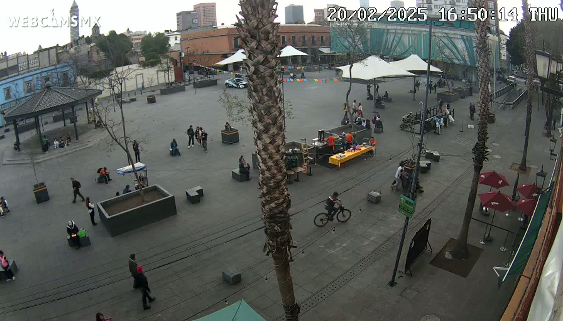 A mostly empty plaza with several people scattered throughout, some sitting at small tables under temporary canopies, under an overcast sky.