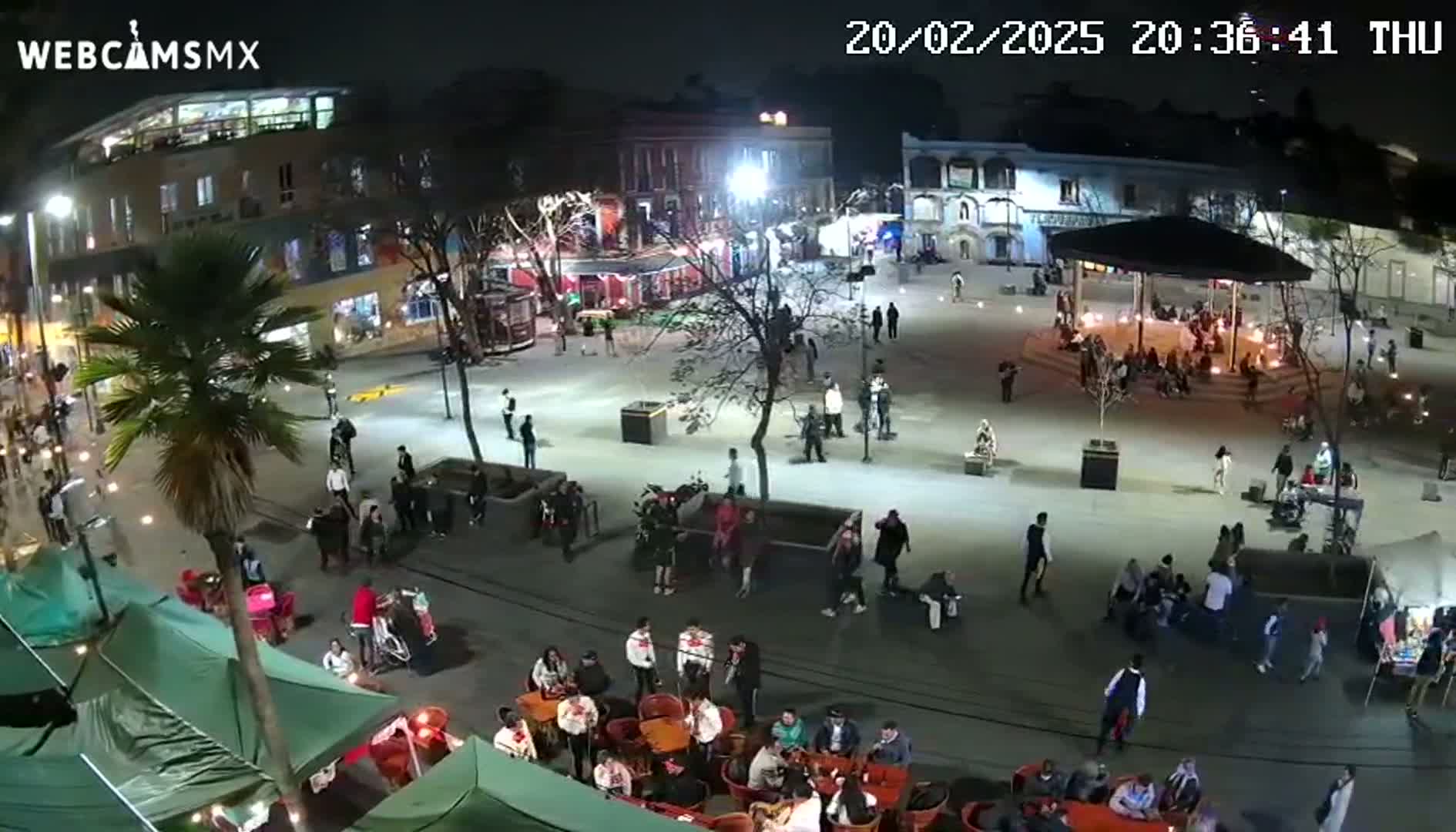 A nighttime aerial view of a town square shows many people gathered around tables at outdoor cafes and in a central gazebo, under clear skies.