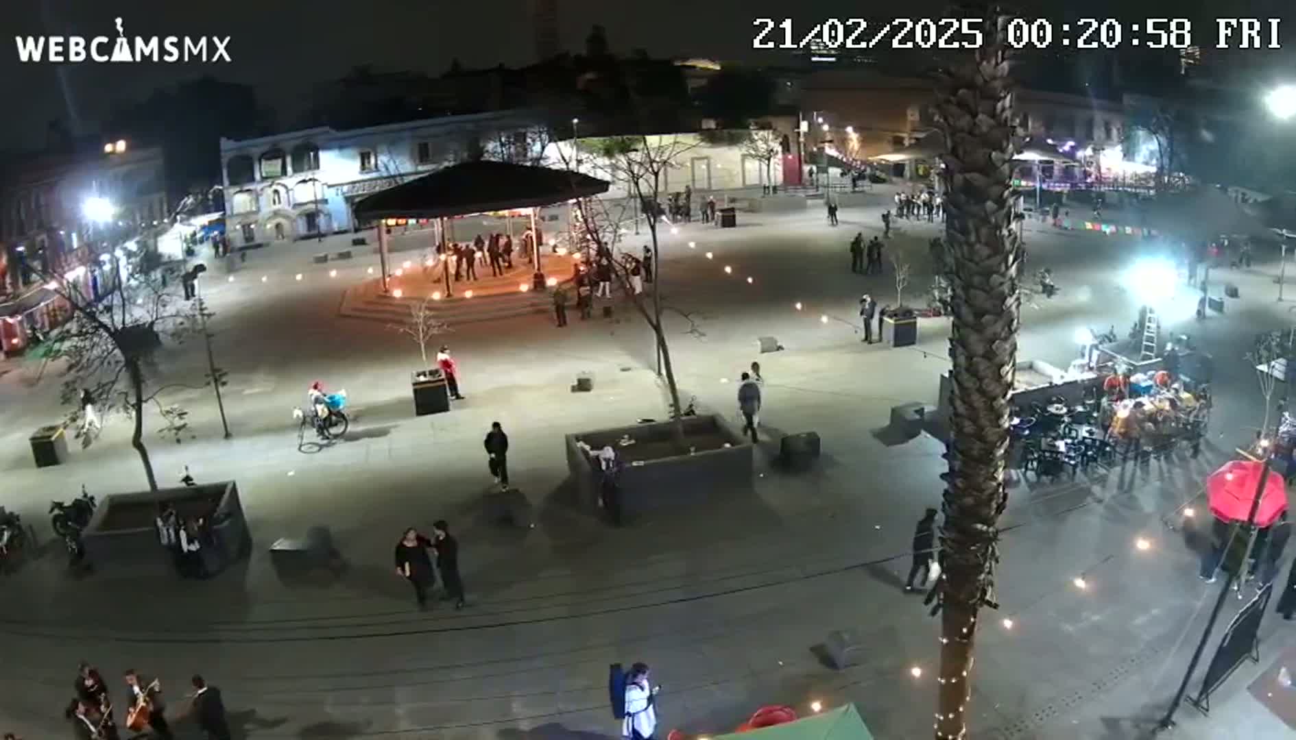 A nighttime aerial view of a town square with people gathered around a gazebo and other areas, illuminated by lights strung across the space.