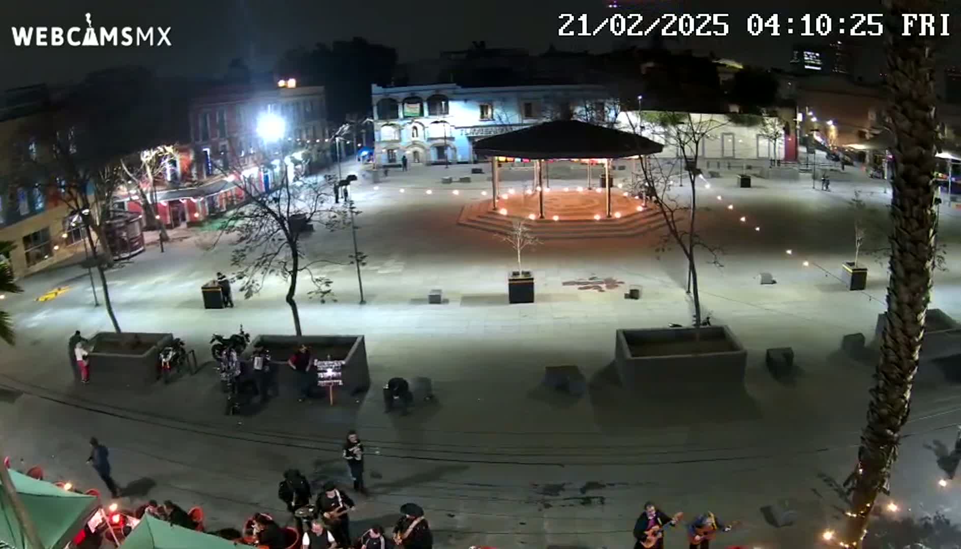 A nighttime view of a plaza with a gazebo in the center, surrounded by buildings and people, under clear skies.