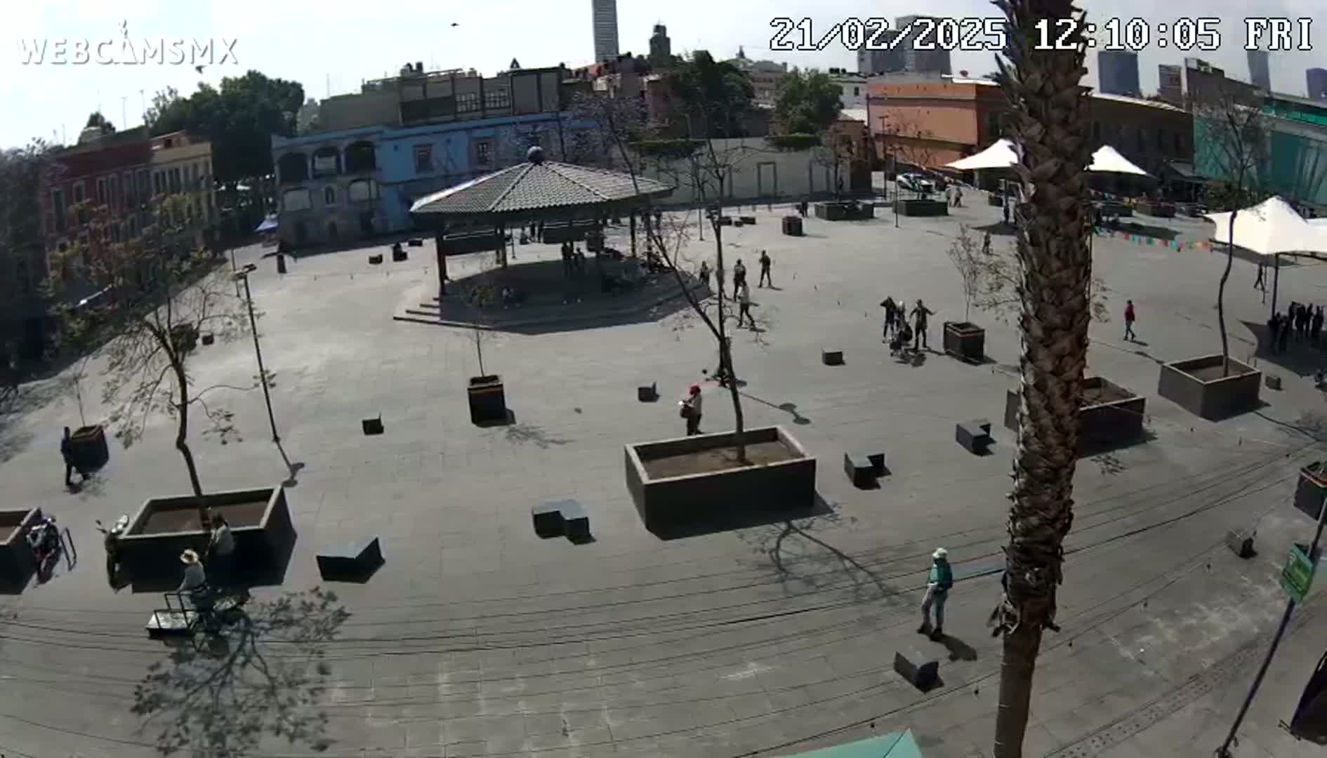 A mostly empty plaza on a sunny day is viewed from above, showing a gazebo, several people, and various trees and planters.