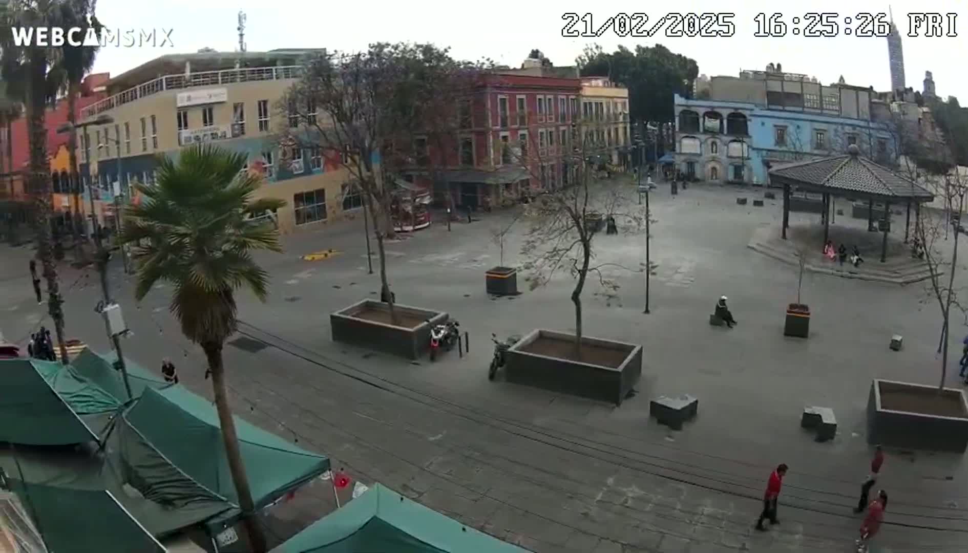 An overcast day reveals a town square with several buildings, a gazebo, some people, and two motorcycles parked near planters.
