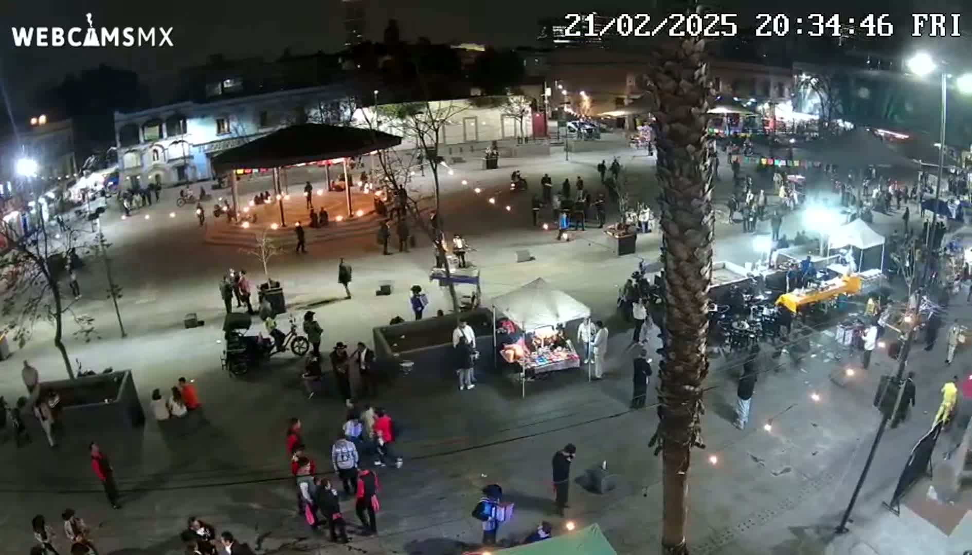 A nighttime aerial view of a town square bustling with people, vendors, and small fires under a clear sky.