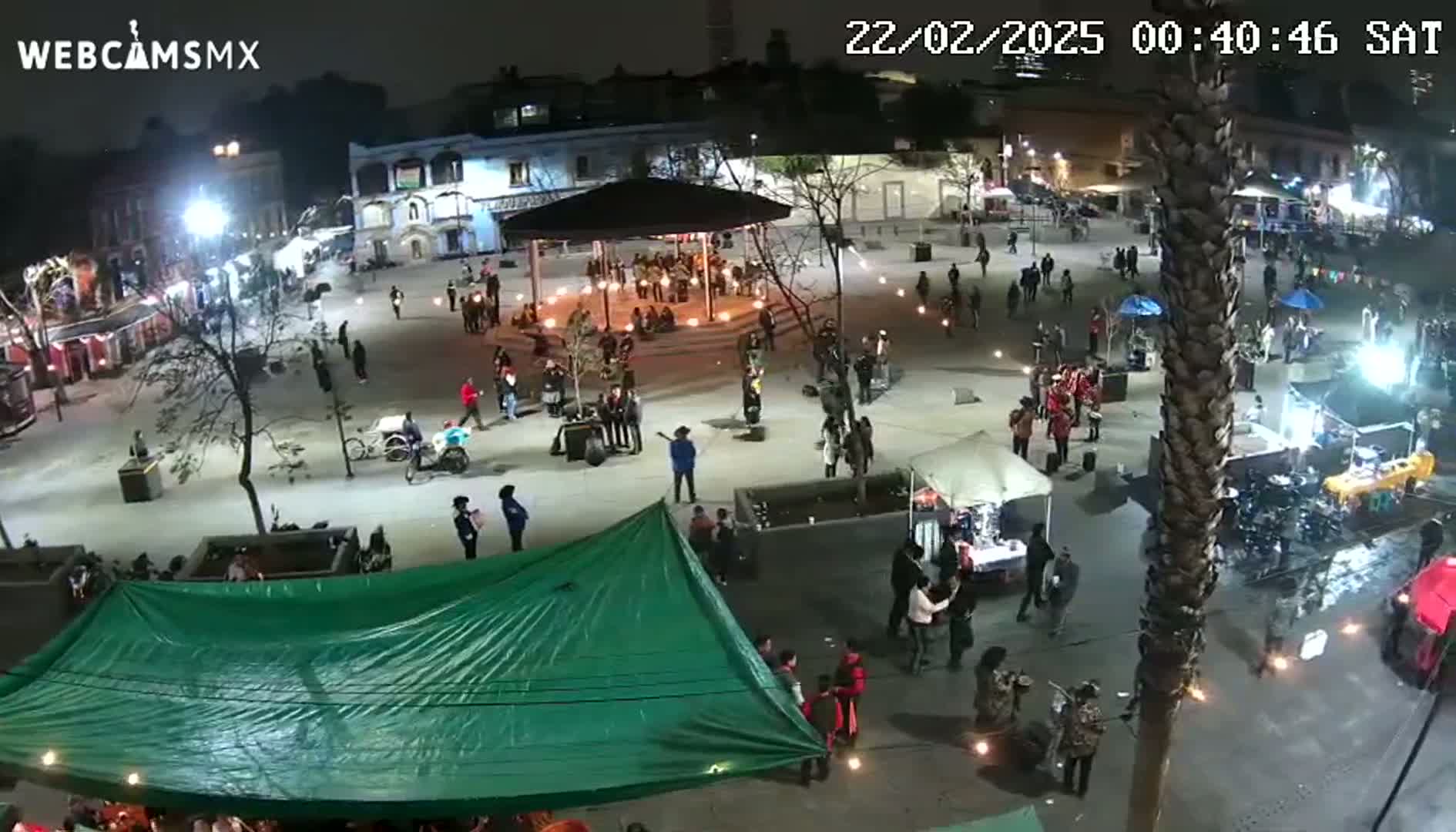 A nighttime, aerial view of a town square with many people gathered around a gazebo and various vendors' stalls, under partly cloudy skies.