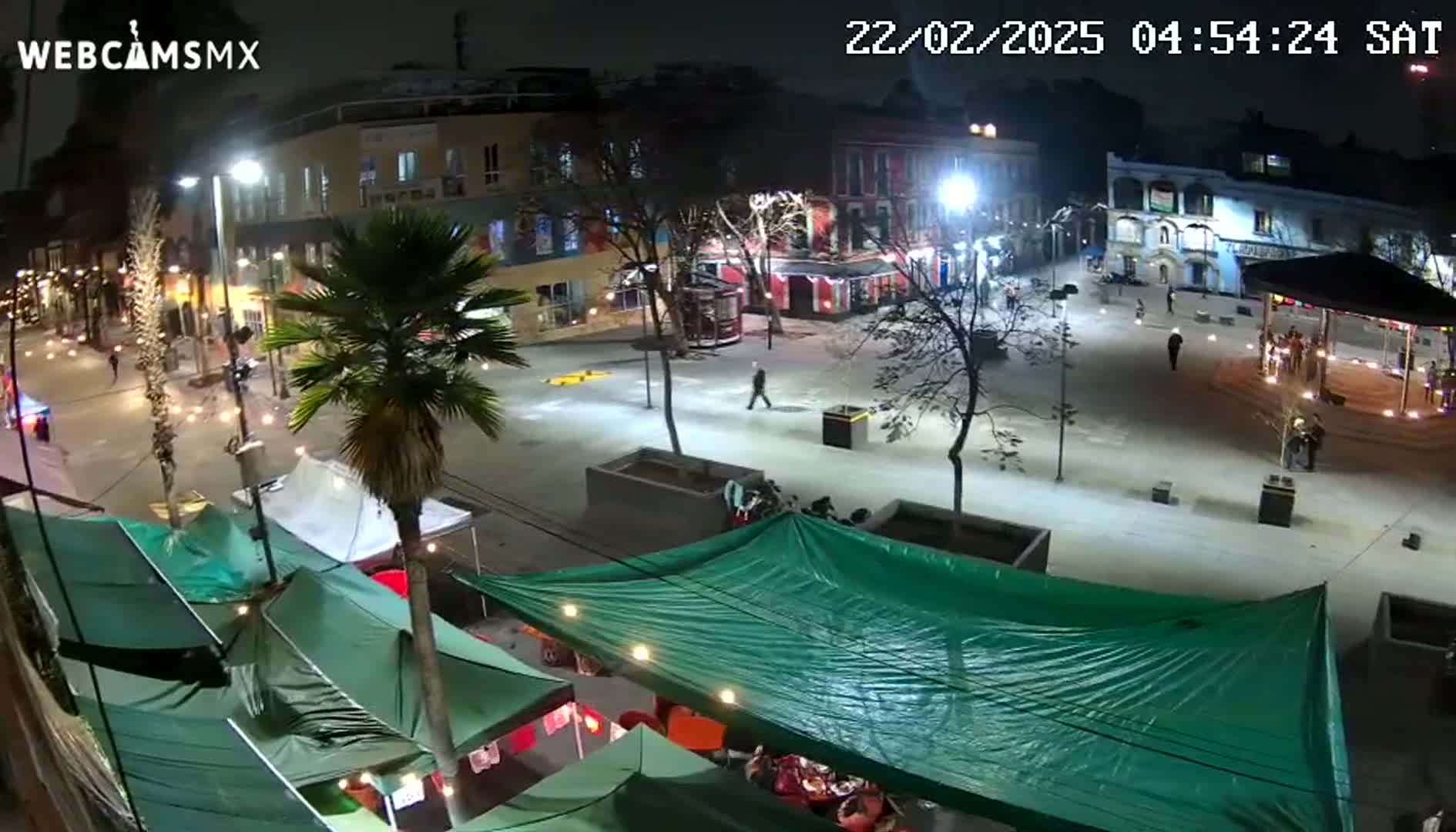 A nighttime, brightly lit plaza scene shows several people walking around, with various tents and buildings surrounding a paved central area.