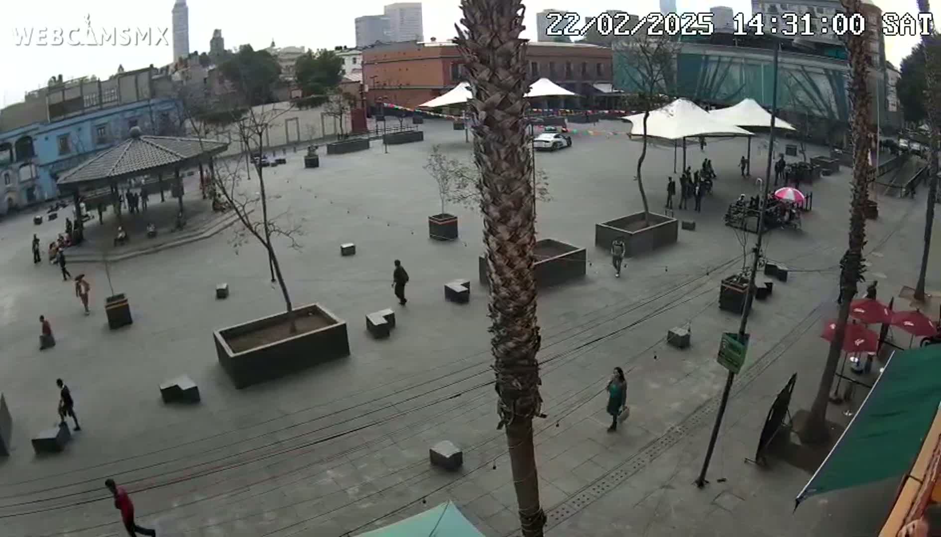 A mostly empty plaza with several people walking around, some sitting under a gazebo, and some under canopies;  the sky is overcast.