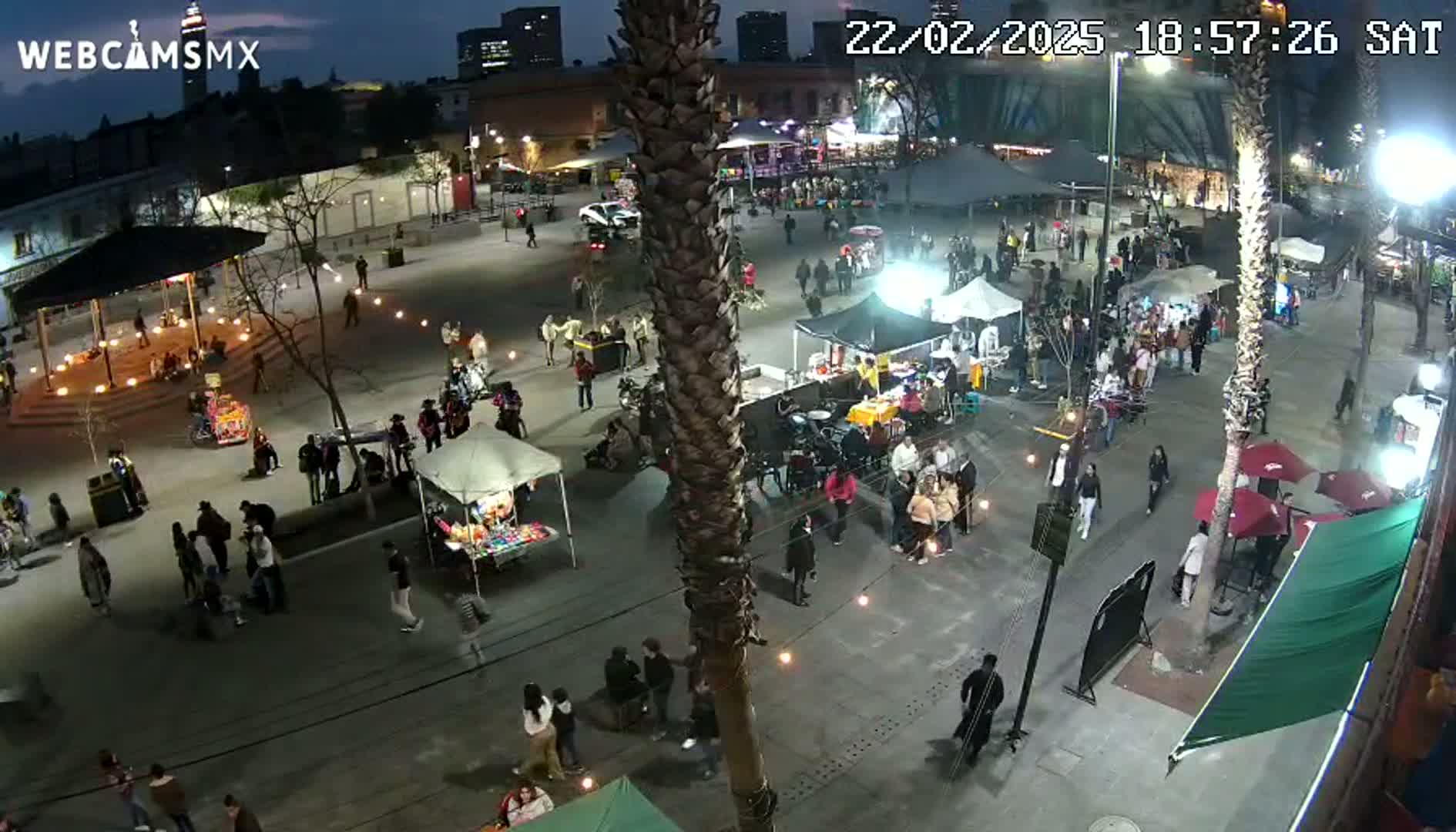 A nighttime aerial view of a bustling town square with numerous people, market stalls, and string lights, under clear skies.