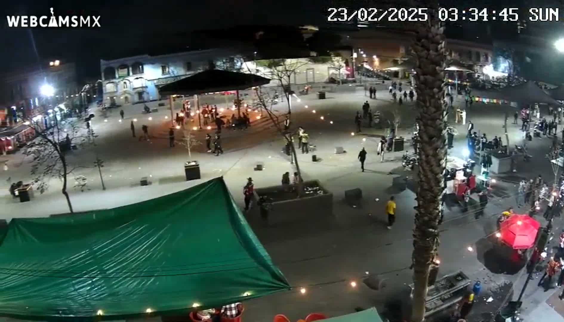 A nighttime aerial view of a town square with people gathered around a gazebo and various food stalls, under clear skies.