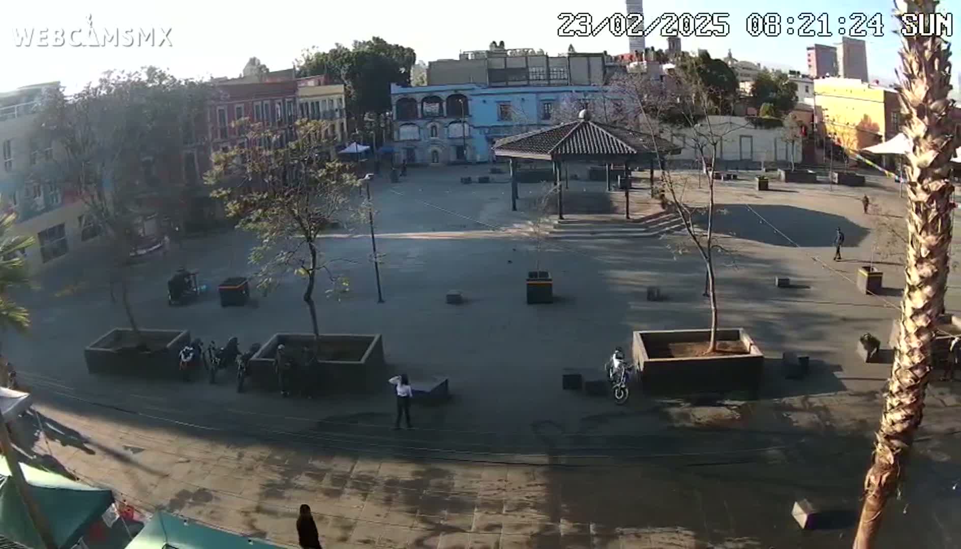 A mostly empty plaza with a gazebo and several buildings surrounding it on a sunny day, with a few people sparsely scattered throughout the area.