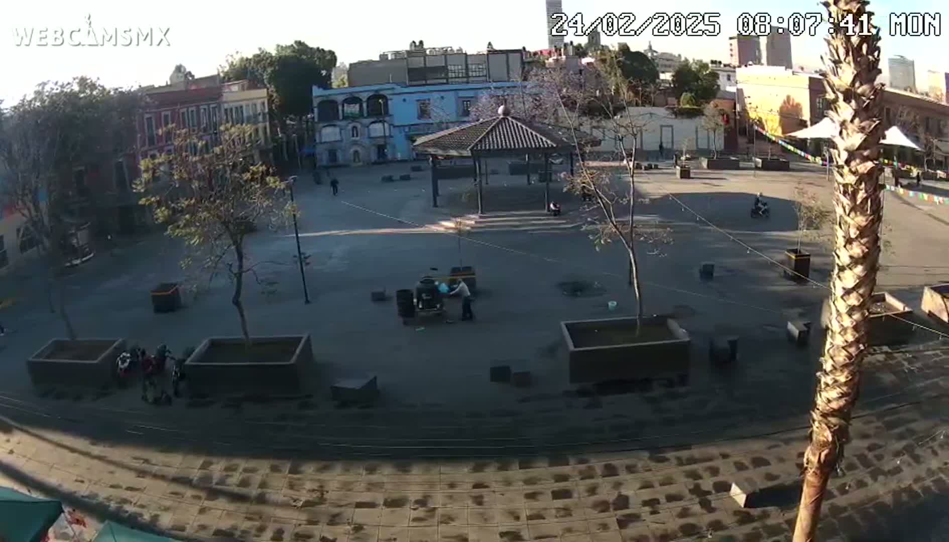 A mostly empty plaza with a gazebo, some people, and several trees, under a sunny sky.