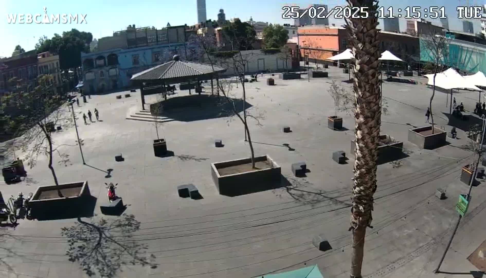 A mostly empty plaza with a gazebo, several trees in planters, and a few pedestrians is visible on a sunny day.