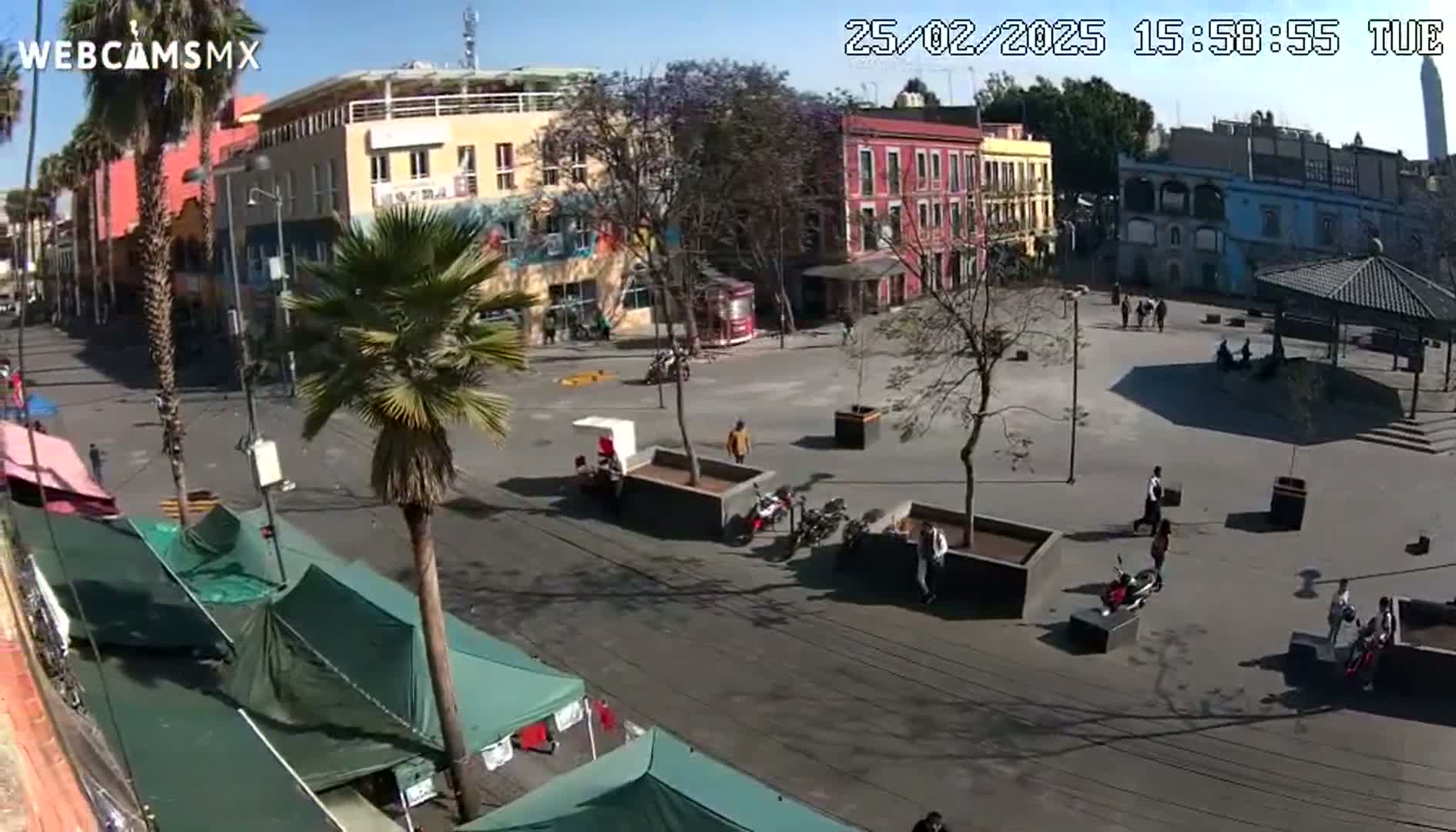 A mostly sunny city square is seen from above, featuring several buildings, a few palm trees, some people walking, and several temporary market stalls.