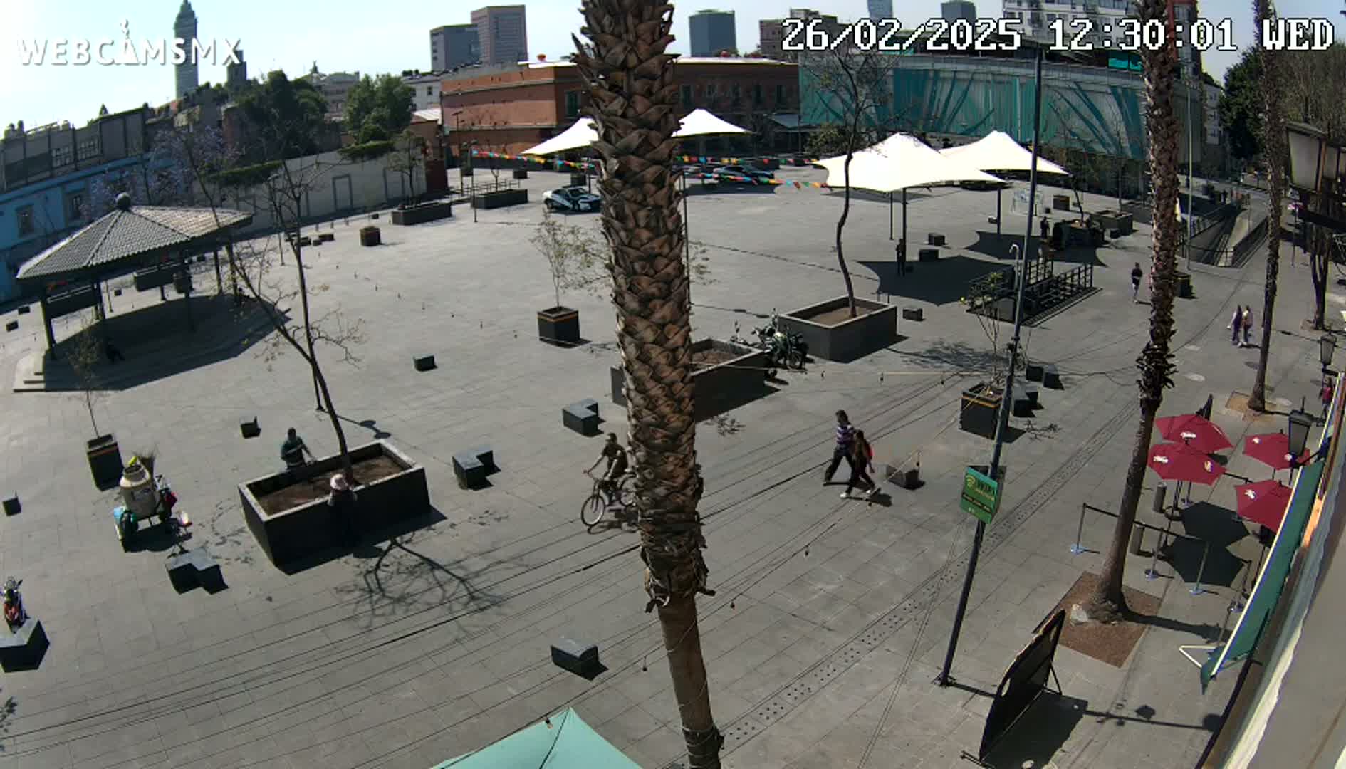 A mostly empty plaza with a few people walking and bicycling, under a sunny sky, is surrounded by buildings and palm trees.