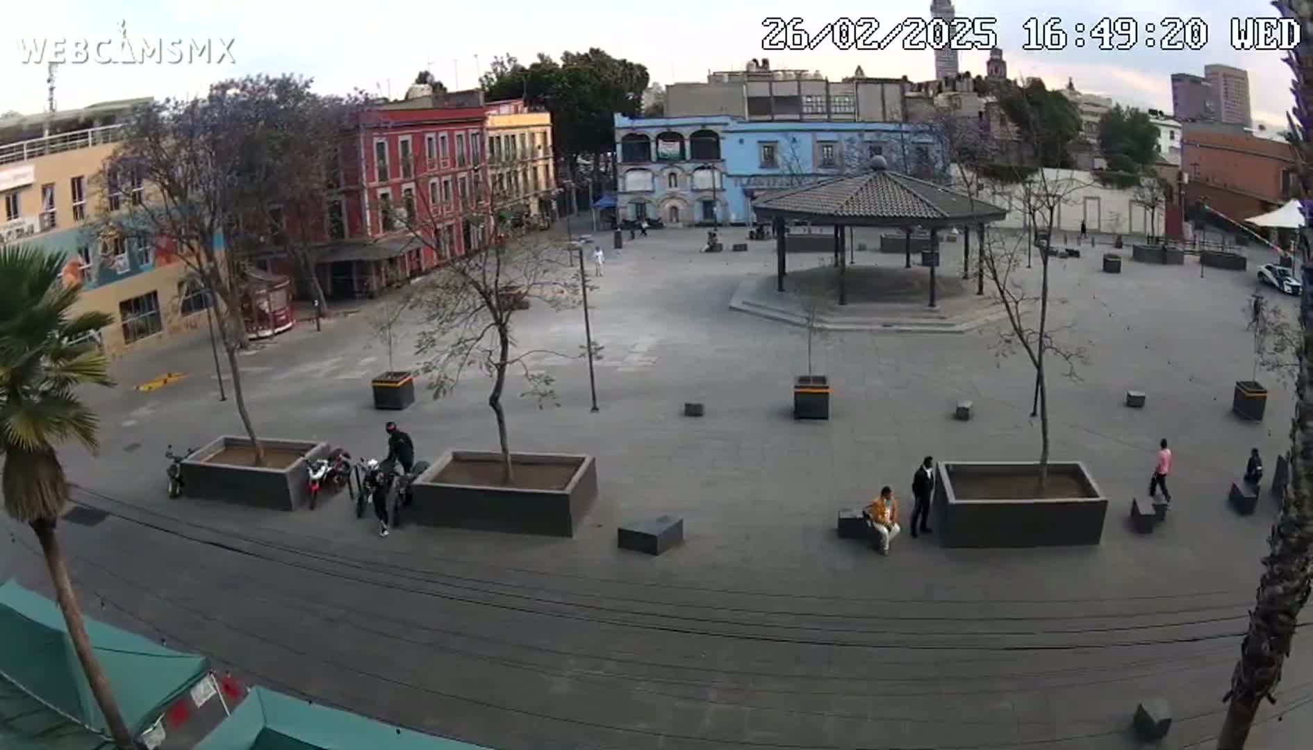 A mostly empty plaza with several buildings surrounding it, a gazebo in the center, and a few people scattered around on a partly cloudy day.