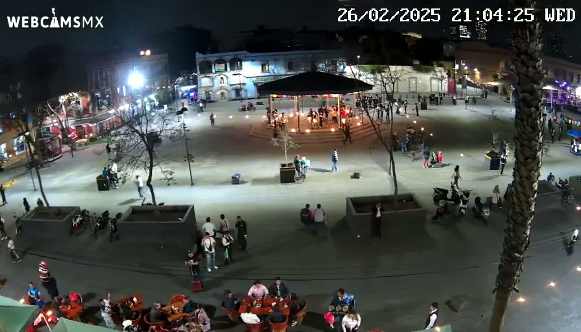 A nighttime, brightly lit plaza with people gathered around tables, a gazebo, and walking throughout the area.