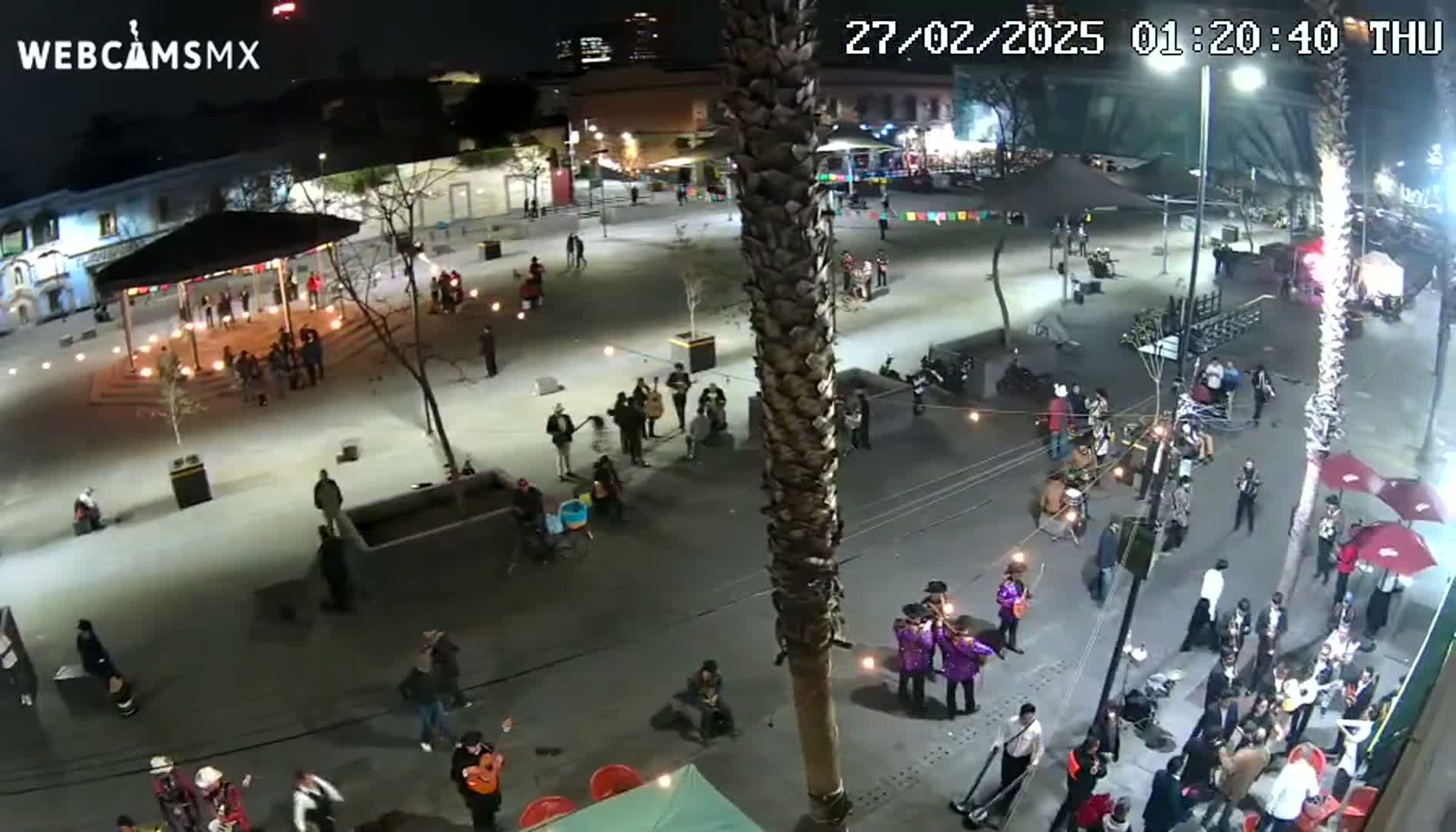 A nighttime aerial view of a plaza with numerous people gathered, some playing music, under illuminated streetlights.