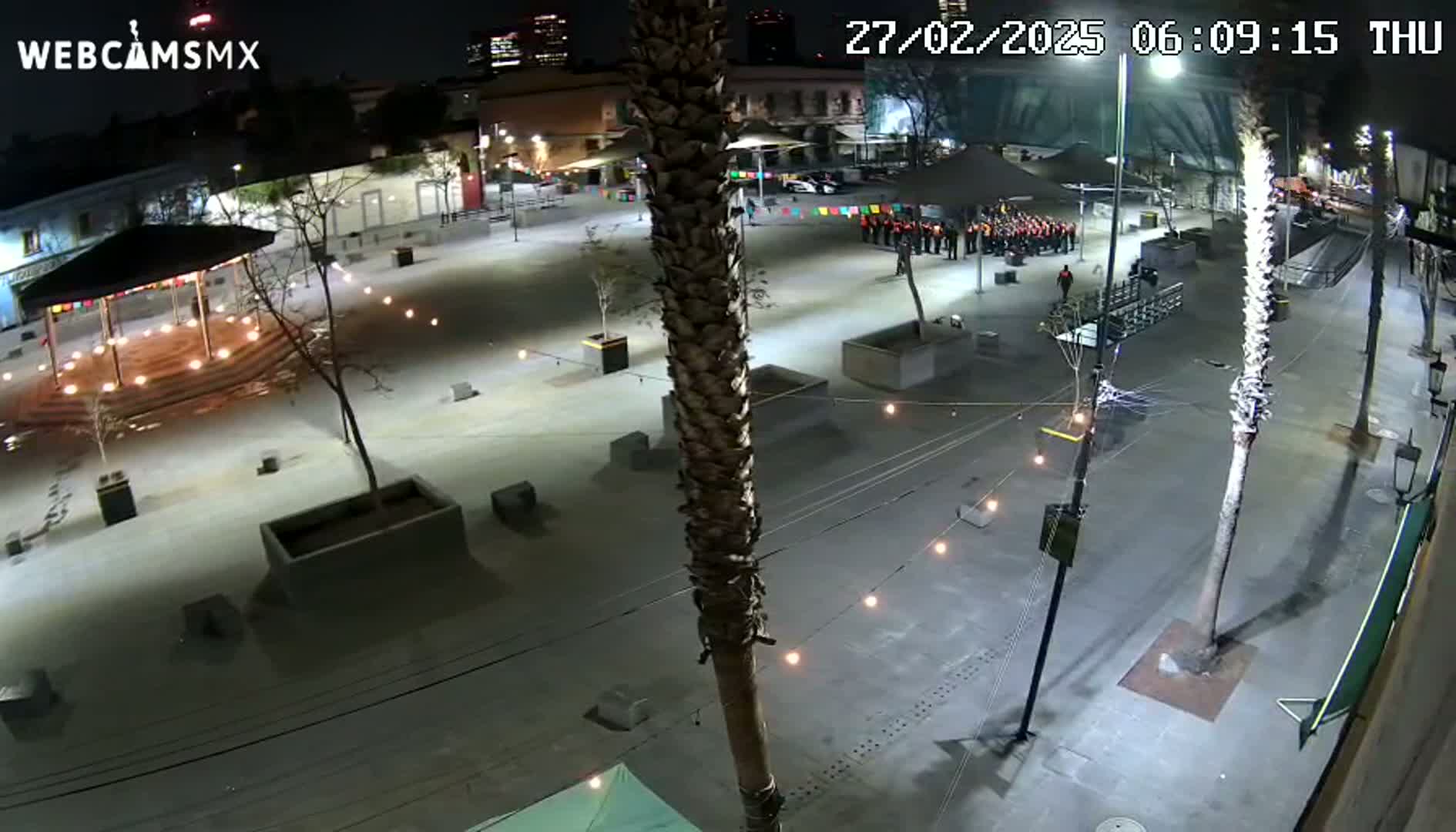 A nighttime view of a plaza with string lights, palm trees, and a small group of people gathered near a pavilion.