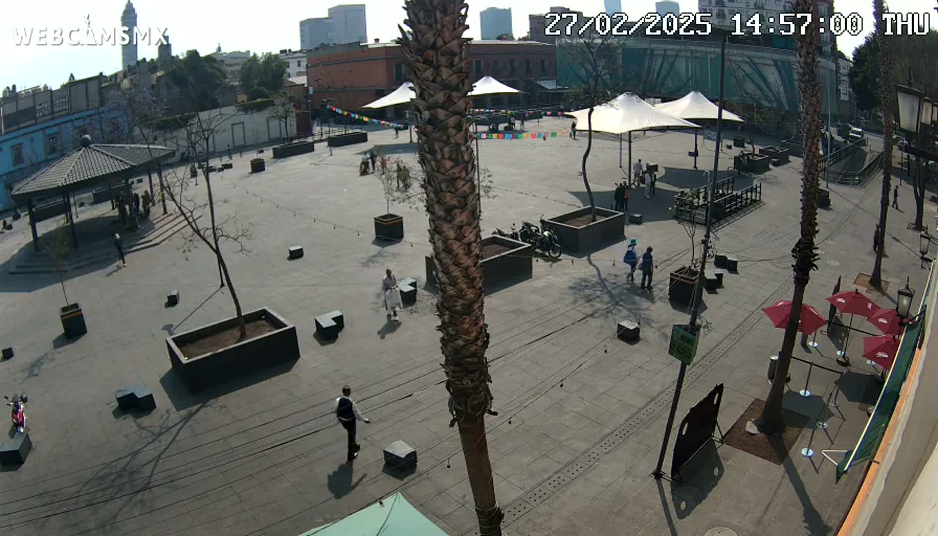 A mostly empty plaza with several people, trees, and seating areas under a sunny sky.