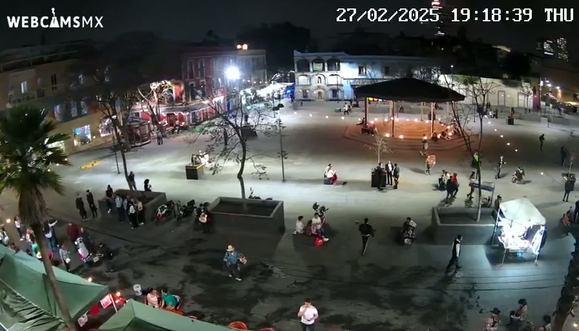 A nighttime, high-angle view shows a town square with people milling about, small vendors, and buildings lining the edges, under clear skies.