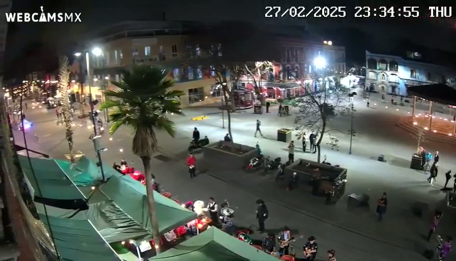 A nighttime, brightly lit town square with pedestrians, parked motorcycles, and a band performing under partially covered market stalls.