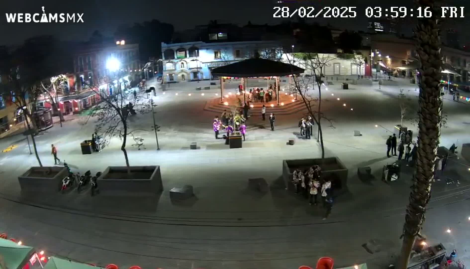 A nighttime aerial view of a plaza with people gathered around a gazebo, illuminated by artificial lights.