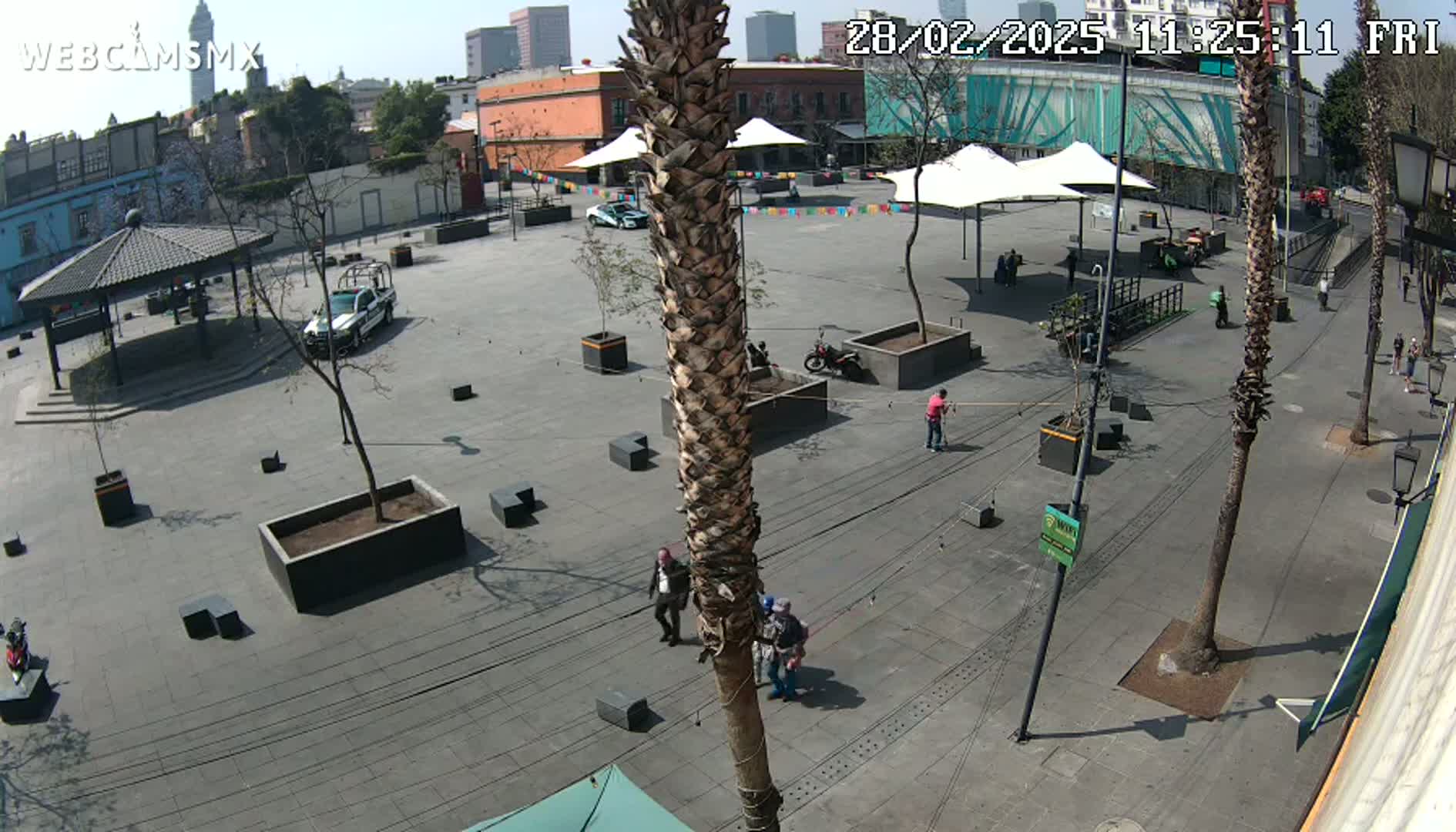 A sunny outdoor plaza scene shows several people walking around, with a police car and a motorcycle parked, and various potted plants and trees scattered throughout.
