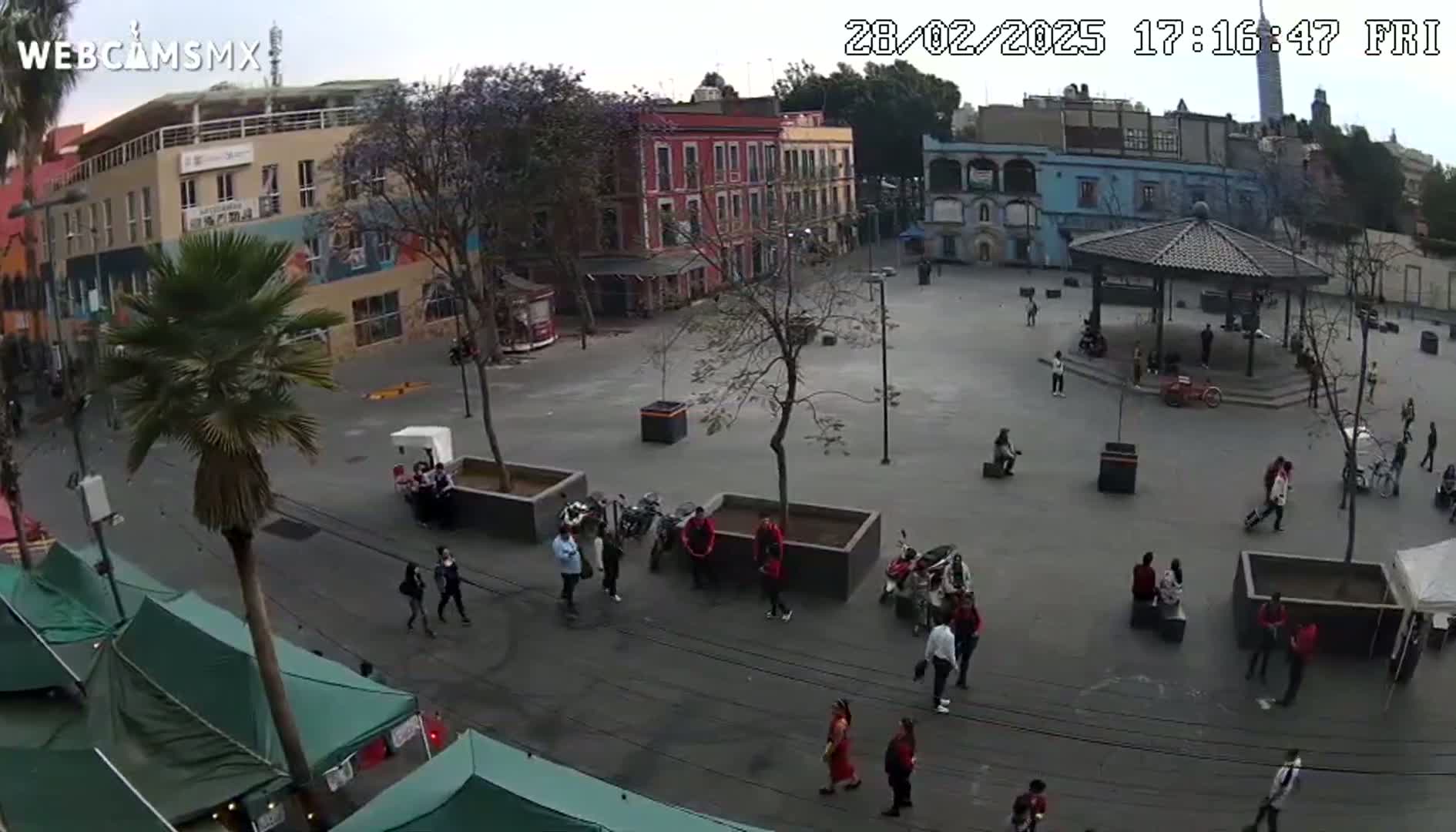 A mostly empty plaza with several people and parked motorcycles is surrounded by colorful buildings under an overcast sky.
