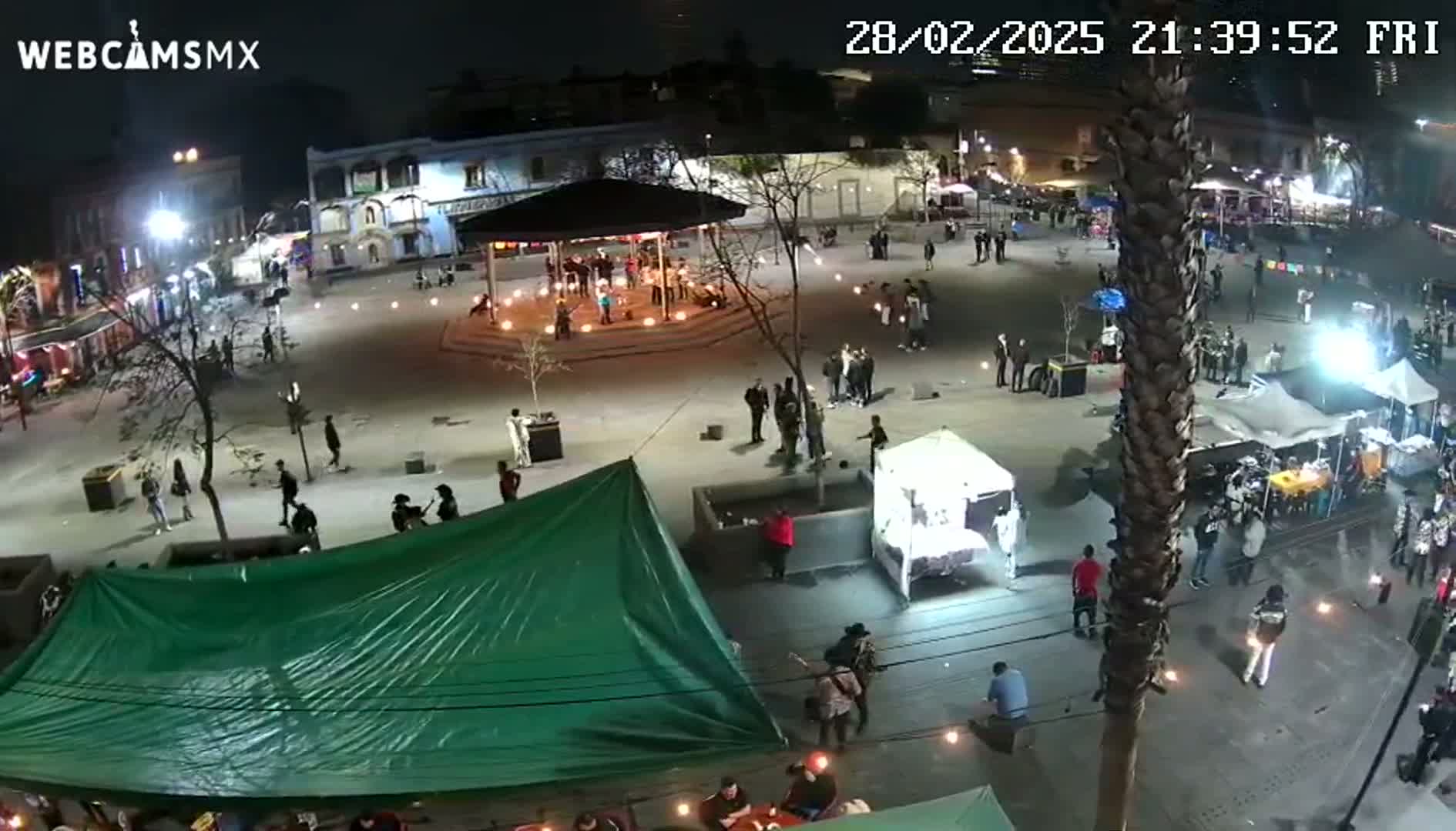 A nighttime outdoor scene shows a town square with people gathered around a gazebo and various small stalls under a clear sky.