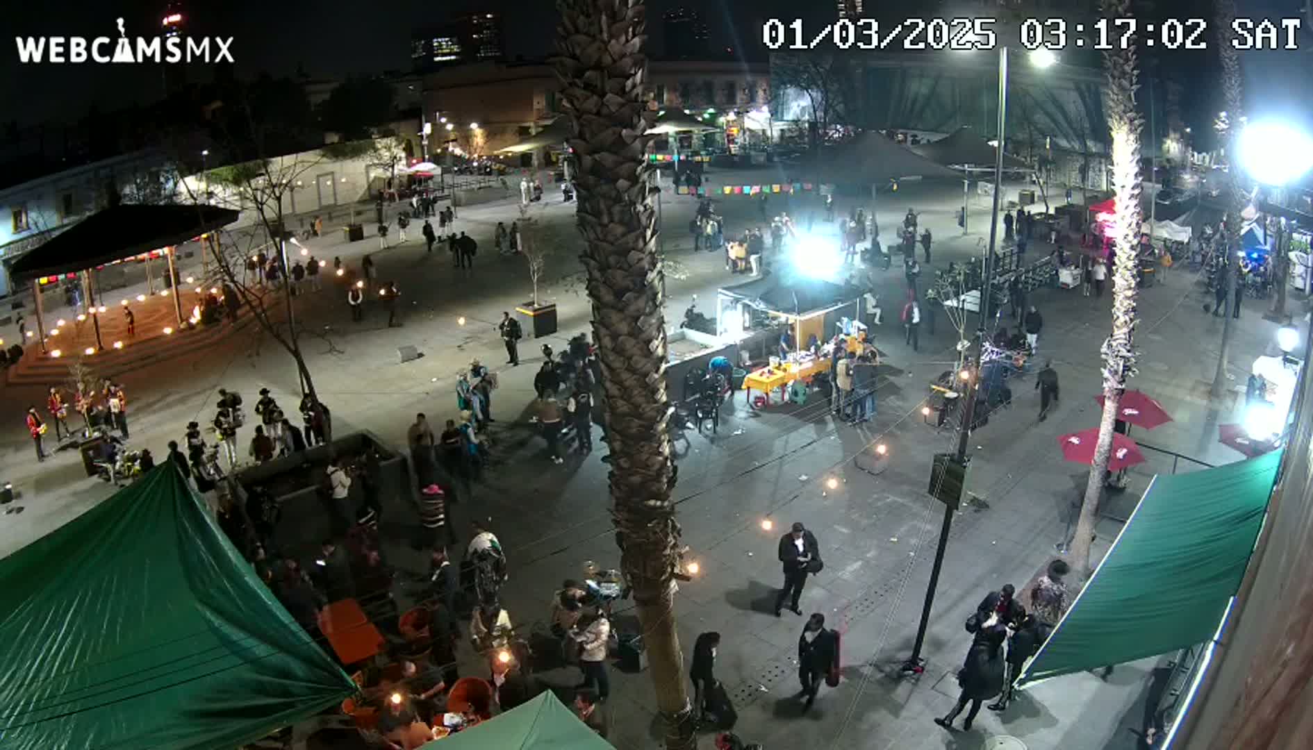 A nighttime, outdoor scene shows a plaza filled with numerous people, some gathered around tables and others milling about, with a band performing near a gazebo, under string lights.