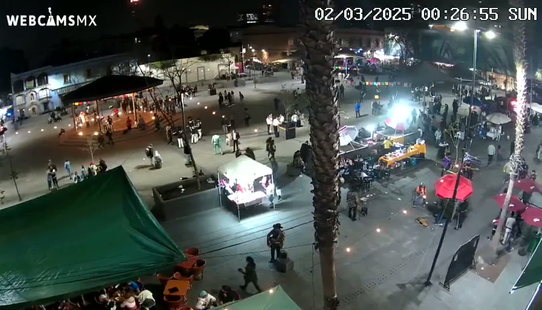 A nighttime aerial view shows a town square filled with people, food stalls, and a gazebo, under clear skies.