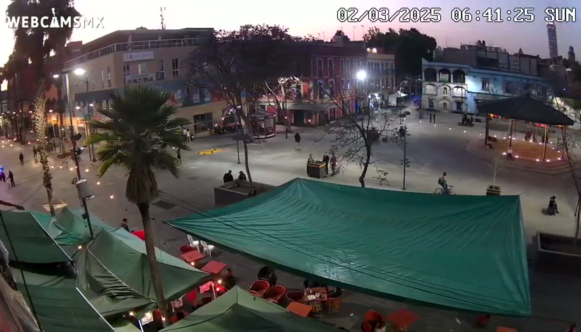 An outdoor plaza at dusk, with several people milling around,  a large green tarp covering an outdoor seating area, and buildings lining the perimeter.