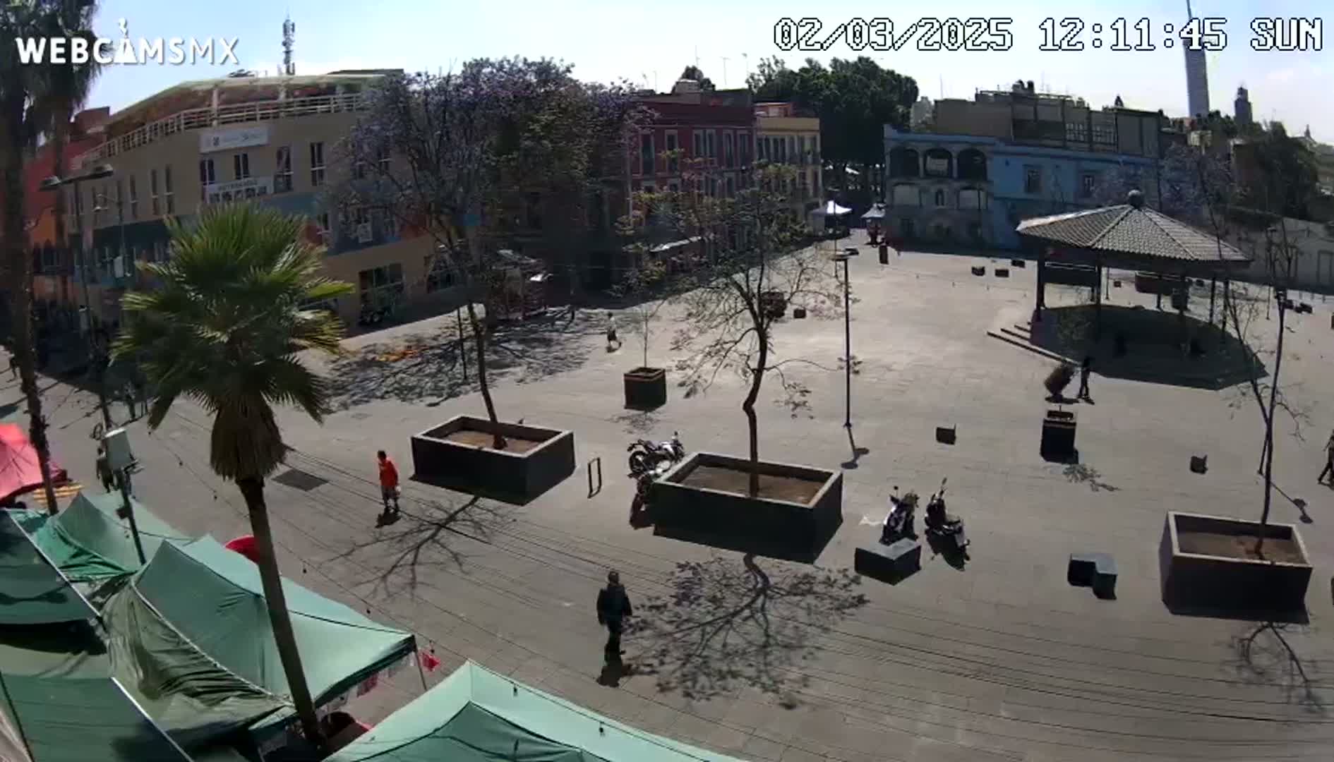 A sunny day reveals a plaza with several buildings, trees, a gazebo, and a few people scattered around.