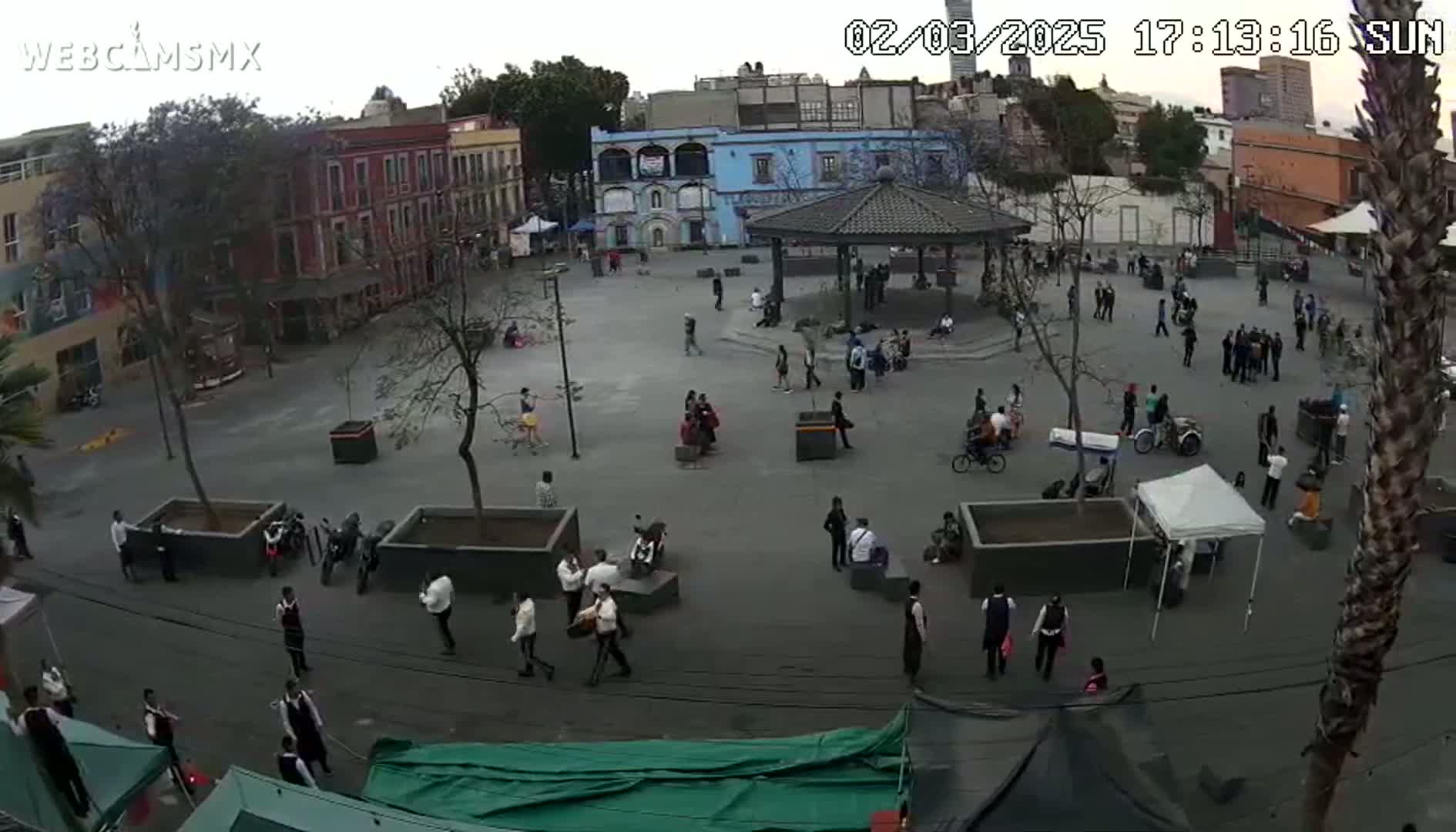 A mostly overcast view of a town square with many people gathered in small groups, some seated under a gazebo, with buildings surrounding the plaza.